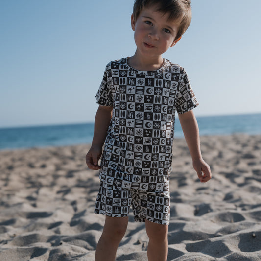 A young child wearing forever french baby’s Bamboo Short Set | Summer Check stands on a sandy beach, their comfy, hypoallergenic pajamas contrasting beautifully with the sea and clear blue sky in the background.