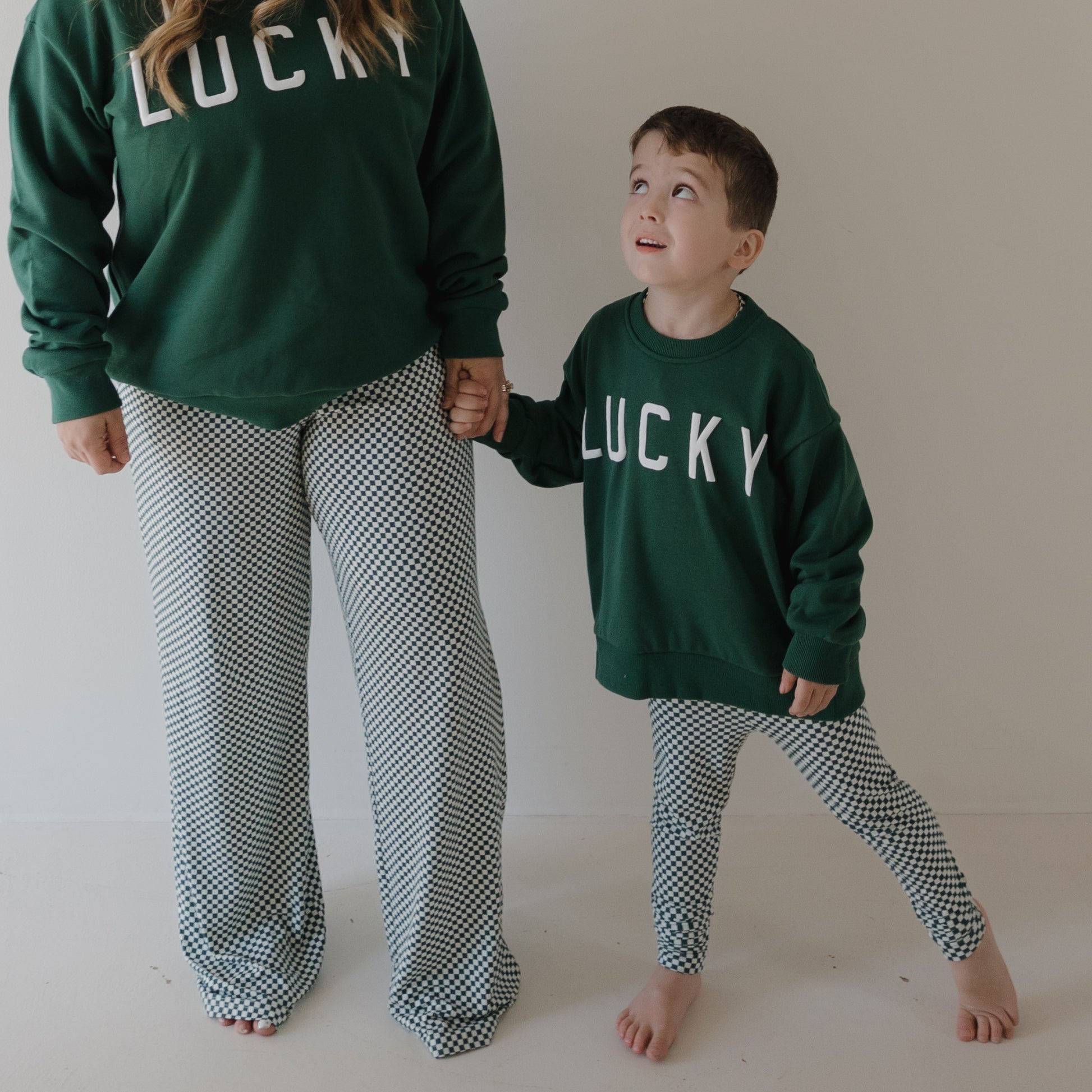 An adult and child stand barefoot against a plain wall, both in forever french baby Bamboo Two Piece Pajamas | Lucky Micro Check sets. The child holds the adult’s hand, looking up while wearing matching green “LUCKY” sweatshirts and checkered pajamas.