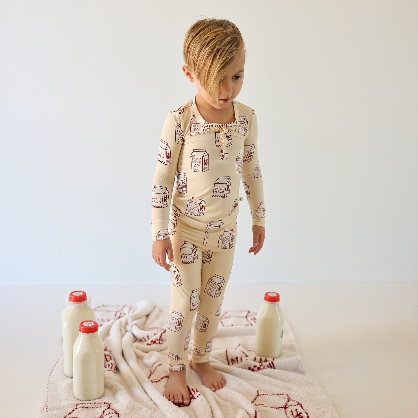A young child stands barefoot on a white towel with red designs, surrounded by three glass milk bottles. They wear forever french baby Bamboo Two Piece Pajamas | Milk—soft, breathable kids’ sleepwear patterned with milk cartons.