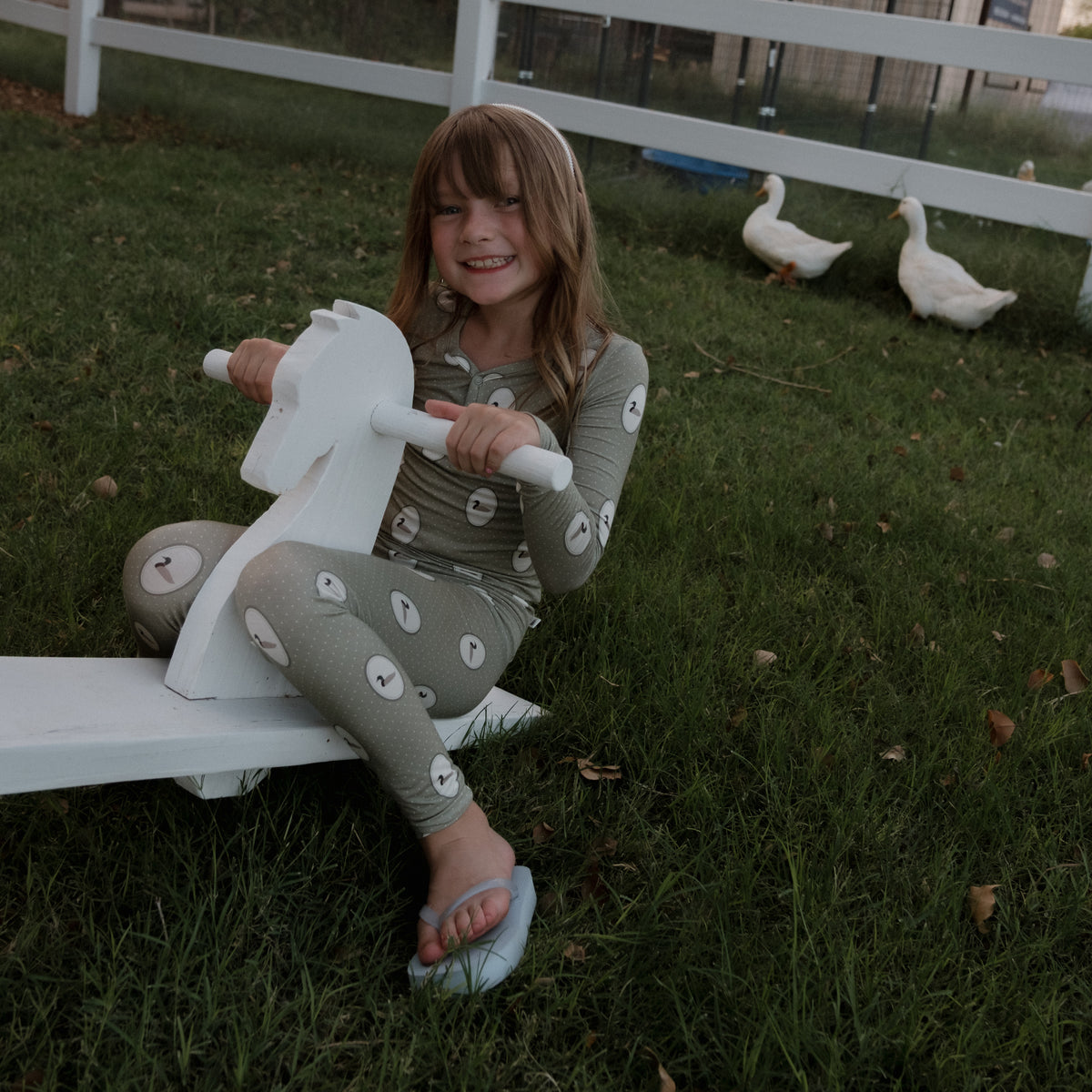 A young girl, smiling at the camera, sits on a white rocking horse atop green grass while wearing forever french baby’s Bamboo Two Piece Pajamas in the Mallard Frames print. In the background, two white ducks walk near a white fence.