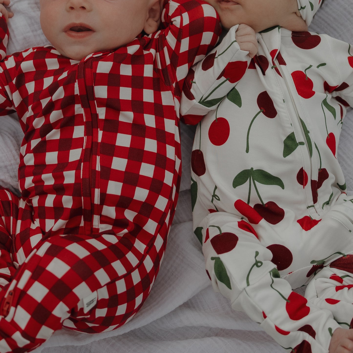 Two babies lie on a white blanket—one in red and white checks, the other in forever french baby’s Bamboo Zip Pajamas | Cherry, featuring breathable bamboo fabric with red cherry prints—both look cozy and cute in their comfy outfits.