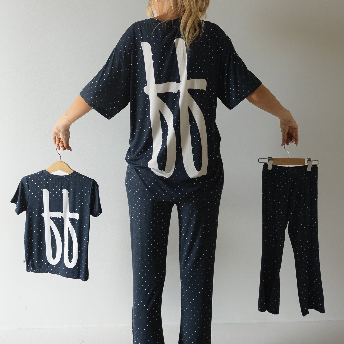 A person faces away, holding the forever french baby Child Short Sleeve Bamboo Lounge Set | Emery for a child and wearing a similar dark polka dot outfit with bold white symbols, all set against a plain light background.