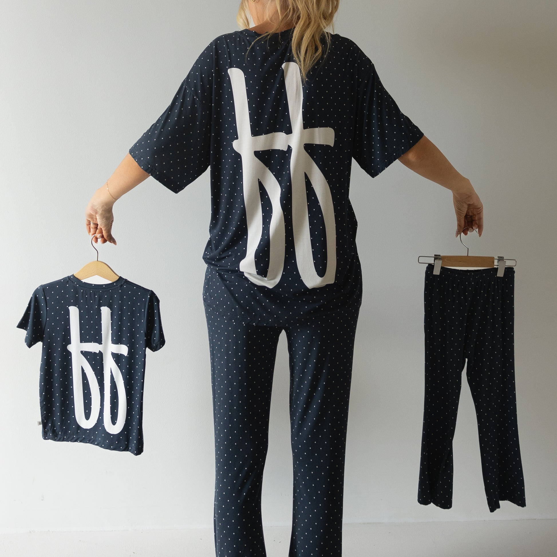 A person faces away, holding the forever french baby Child Short Sleeve Bamboo Lounge Set | Emery for a child and wearing a similar dark polka dot outfit with bold white symbols, all set against a plain light background.