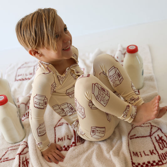 A young child with short, light brown hair sits on a towel with milk carton designs, wearing forever french baby's Bamboo Two Piece Pajamas | Milk. Three glass bottles of milk are placed around them.