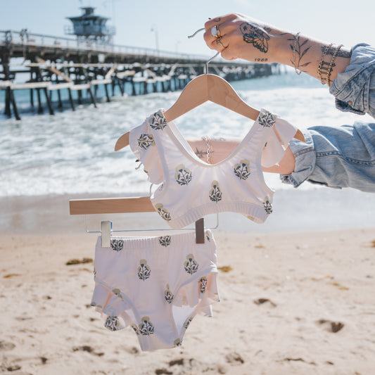 A hand-tattooed person holds up a forever french baby Girl’s Two Piece Swimsuit | Marigold on hangers at a sandy beach, with the pier and ocean waves in the background.