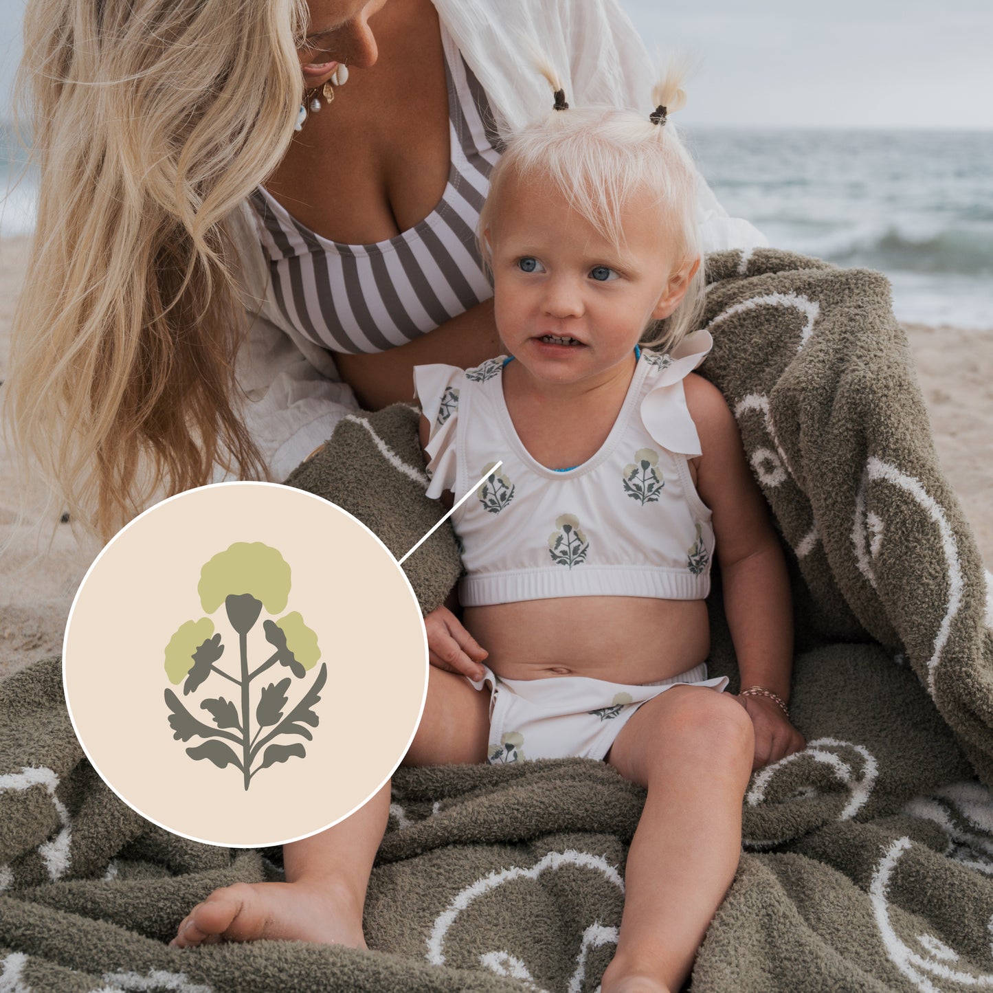 A young girl in the forever french baby Girl's Two Piece Swimsuit | Marigold sits on a beach blanket with an adult. An inset highlights the floral design, showcasing stylish swimwear for kids. Ocean waves can be seen in the background.