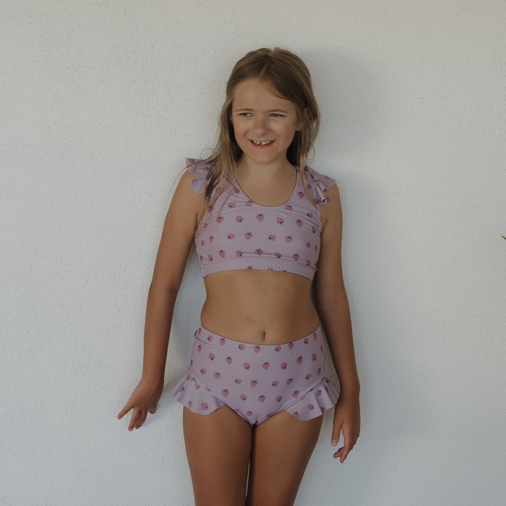 A young girl stands against a plain white wall, smiling and looking to the side, dressed in the forever french baby Girl's Two Piece Swimsuit | Matilda— a light purple set with ruffle details and a subtle pattern.