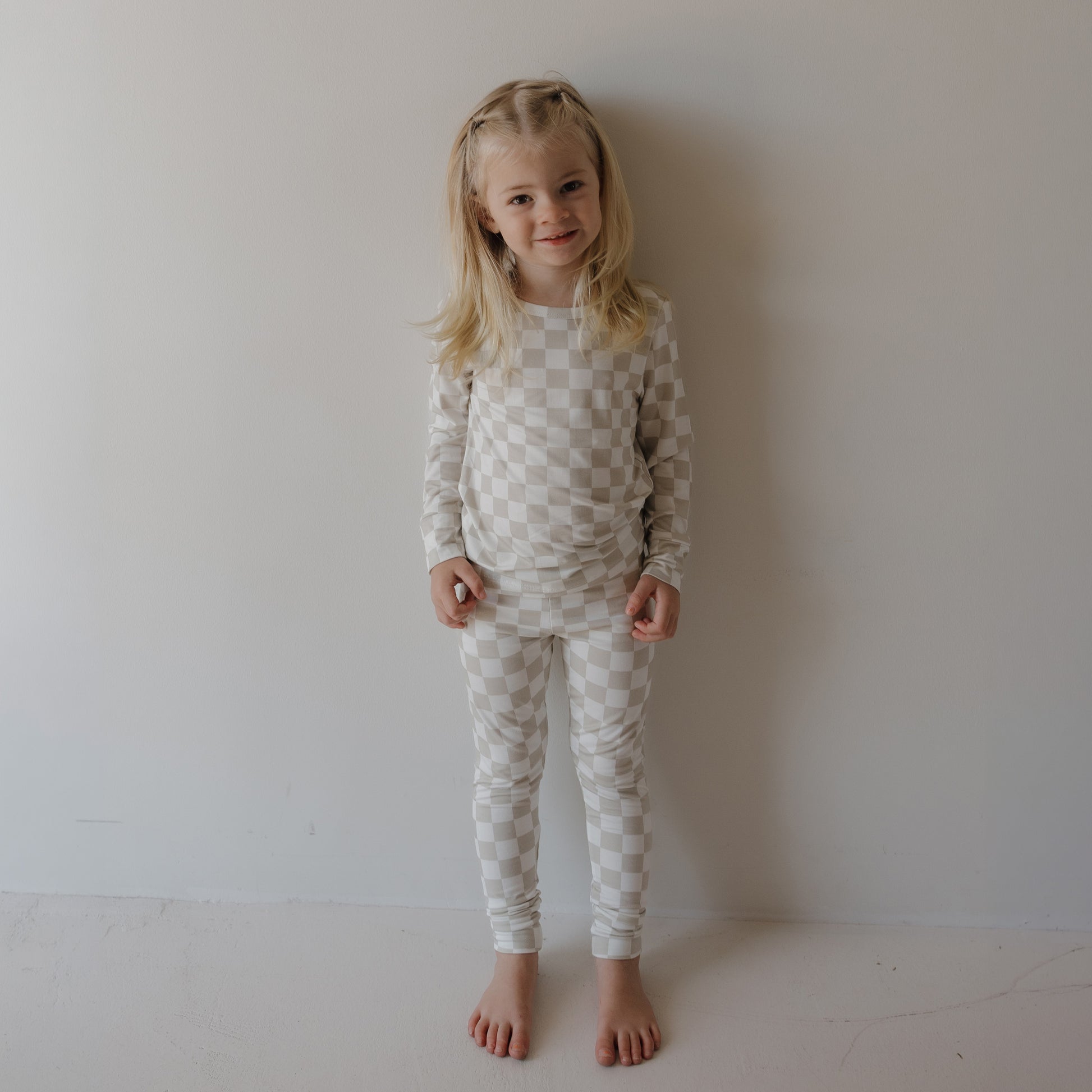 A young child with long blonde hair, barefoot against a plain light wall, smiles gently while wearing the forever french baby Child Sleepy Time Set | Checkerboard in beige and white.