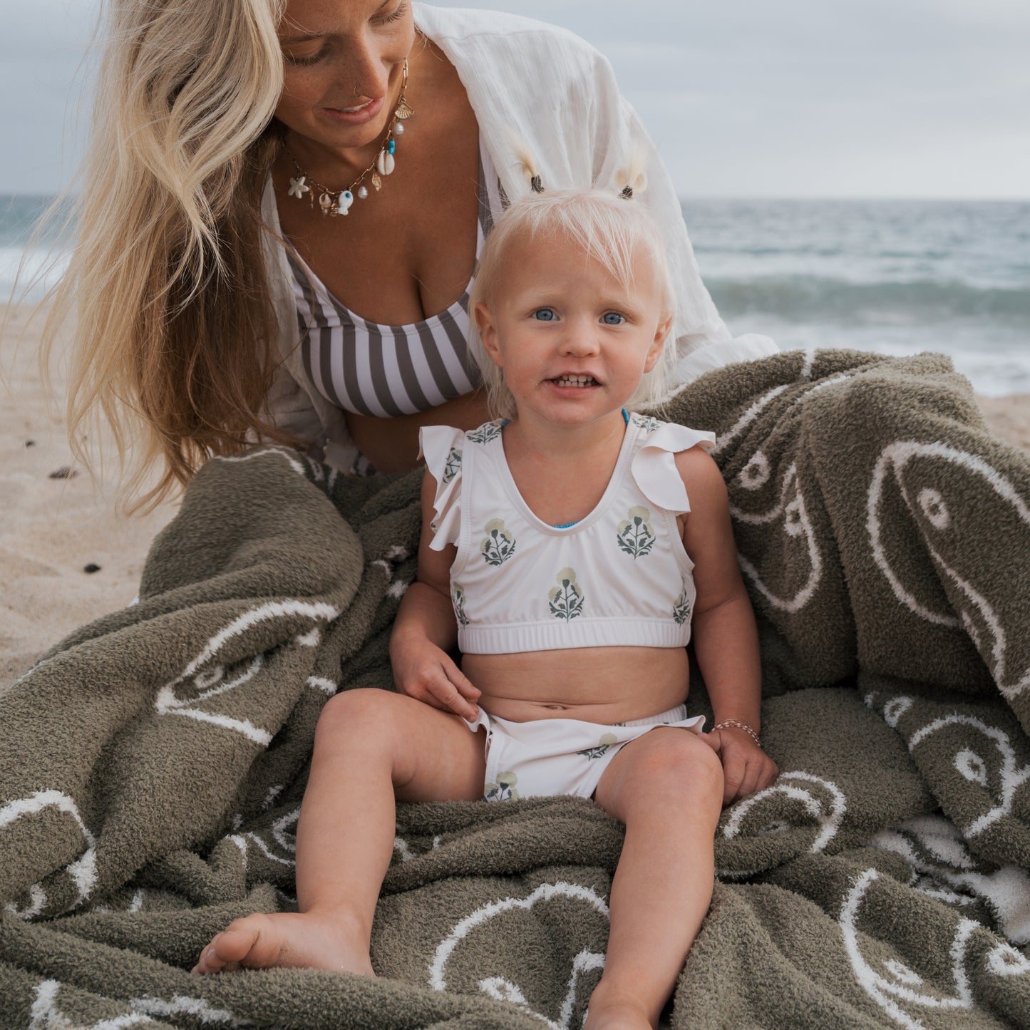 A woman and a young girl sit on the beach, wrapped in a green blanket. The smiling girl wears a forever french baby Girl's Two Piece Swimsuit | Marigold, with the ocean providing a peaceful backdrop.