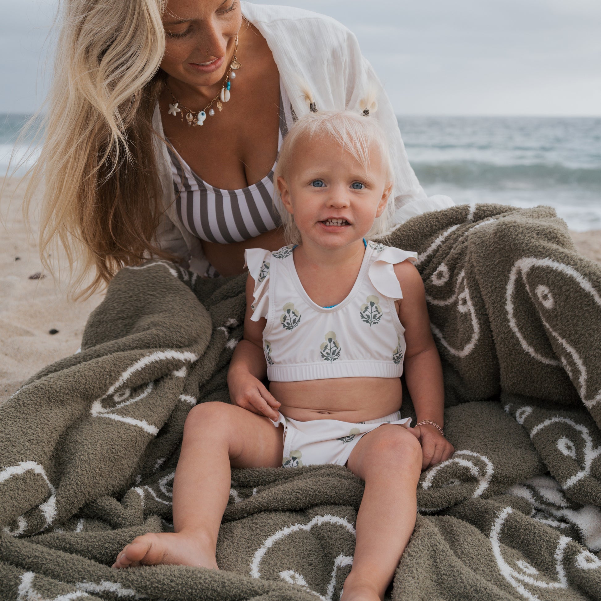 A woman and a young girl sit on the beach, wrapped in a green blanket. The smiling girl wears a forever french baby Girl's Two Piece Swimsuit | Marigold, with the ocean providing a peaceful backdrop.