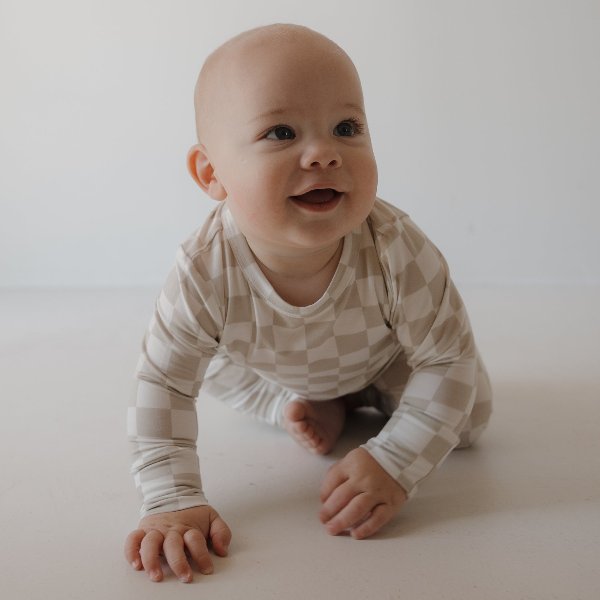 A smiling baby crawls on a white floor, wearing the forever french baby Child Sleepy Time Set | Checkerboard, featuring beige and white checks, against a plain light background.