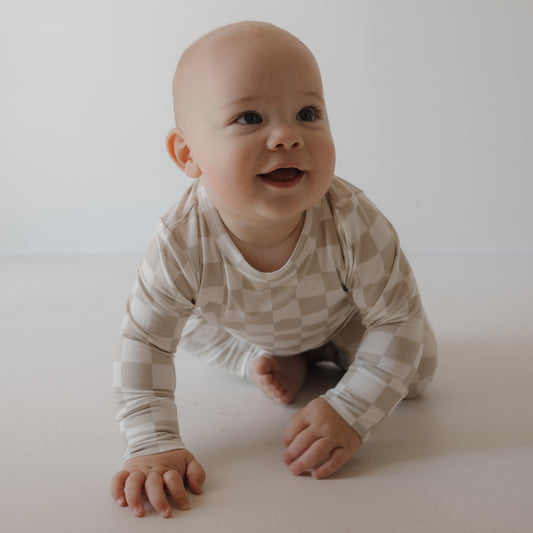 A smiling baby crawls on a white floor, wearing the forever french baby Child Sleepy Time Set | Checkerboard, featuring beige and white checks, against a plain light background.