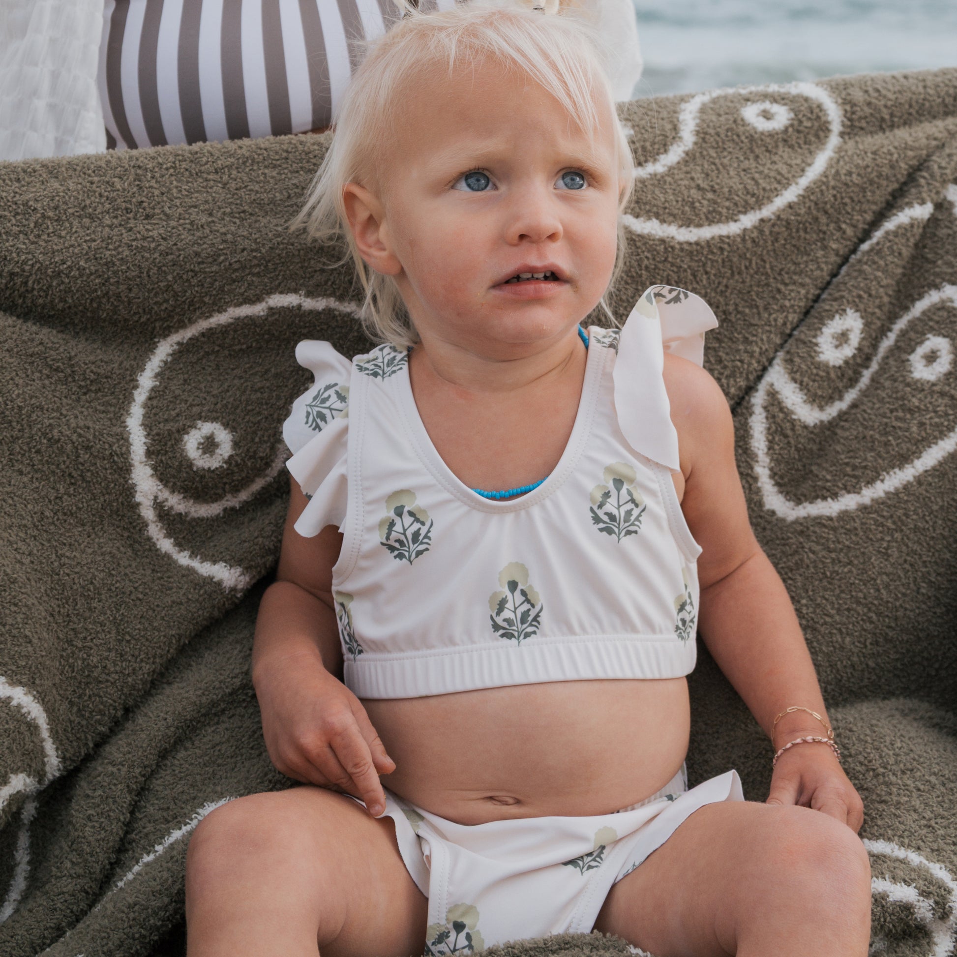A young blonde child sits wrapped in a patterned blanket, wearing the forever french baby Girl's Two Piece Swimsuit | Marigold, featuring floral designs. She looks up with a concerned expression against a softly blurred outdoor backdrop.