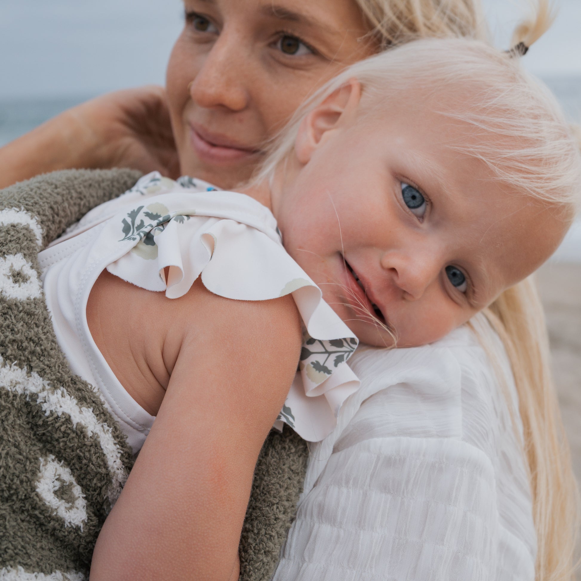 A young woman in a two-piece swimsuit holds a small blonde child wrapped in a green and white towel at the beach. The child, cozy in her forever french baby Girl's Two Piece Swimsuit | Marigold, rests contentedly on the woman's shoulder.