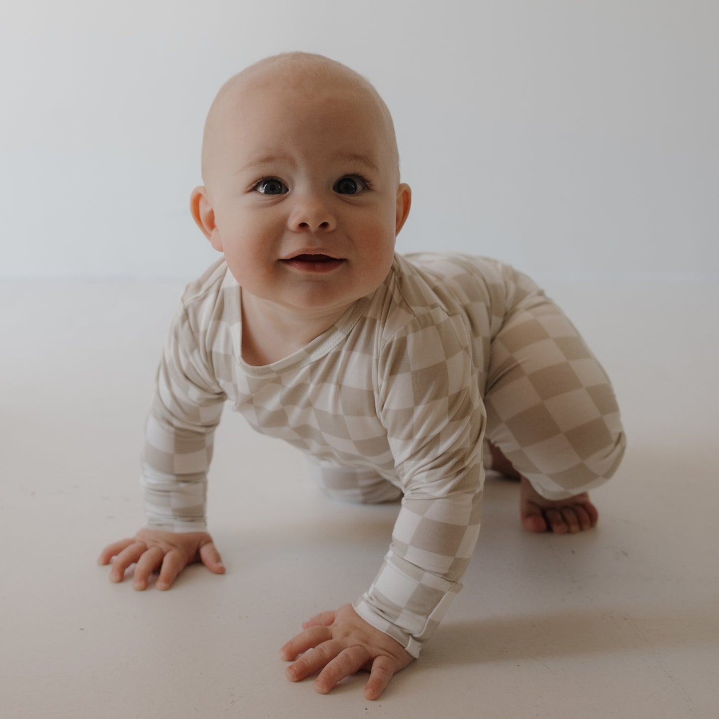 A smiling baby in the forever french baby Child Sleepy Time Set | Checkerboard crawls on a white floor against a plain white background.