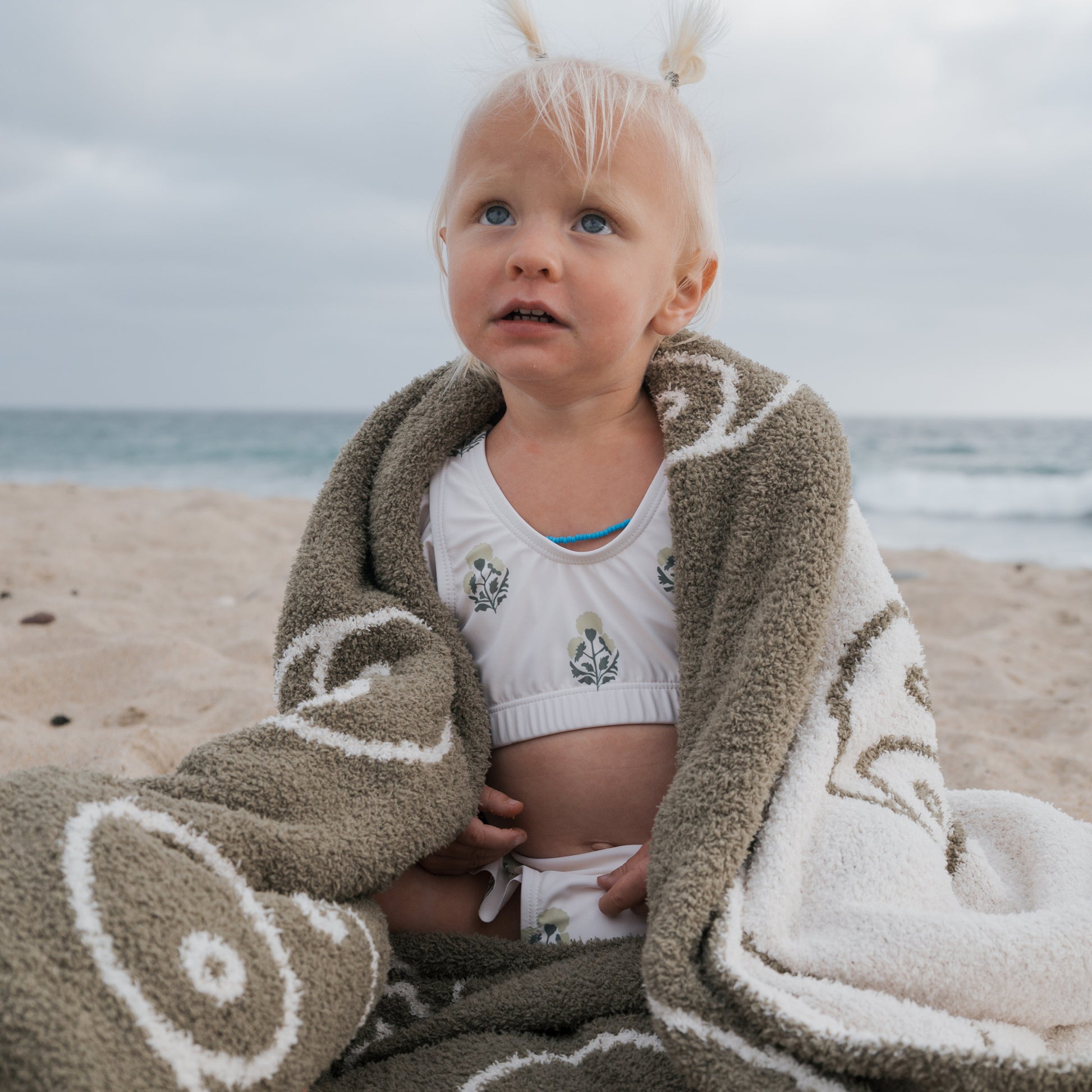 A young child with blonde hair sits on a sandy beach, wrapped in a brown and white towel, wearing the forever french baby Girl's Two Piece Swimsuit in Marigold. The ocean and cloudy sky create a peaceful backdrop.