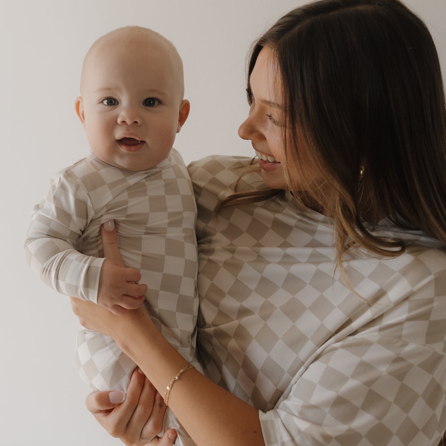 A smiling woman holds a baby as they wear matching forever french baby Women's Boxy Tee Lounge Sets in a beige and white checkerboard pattern, standing against a plain light background.