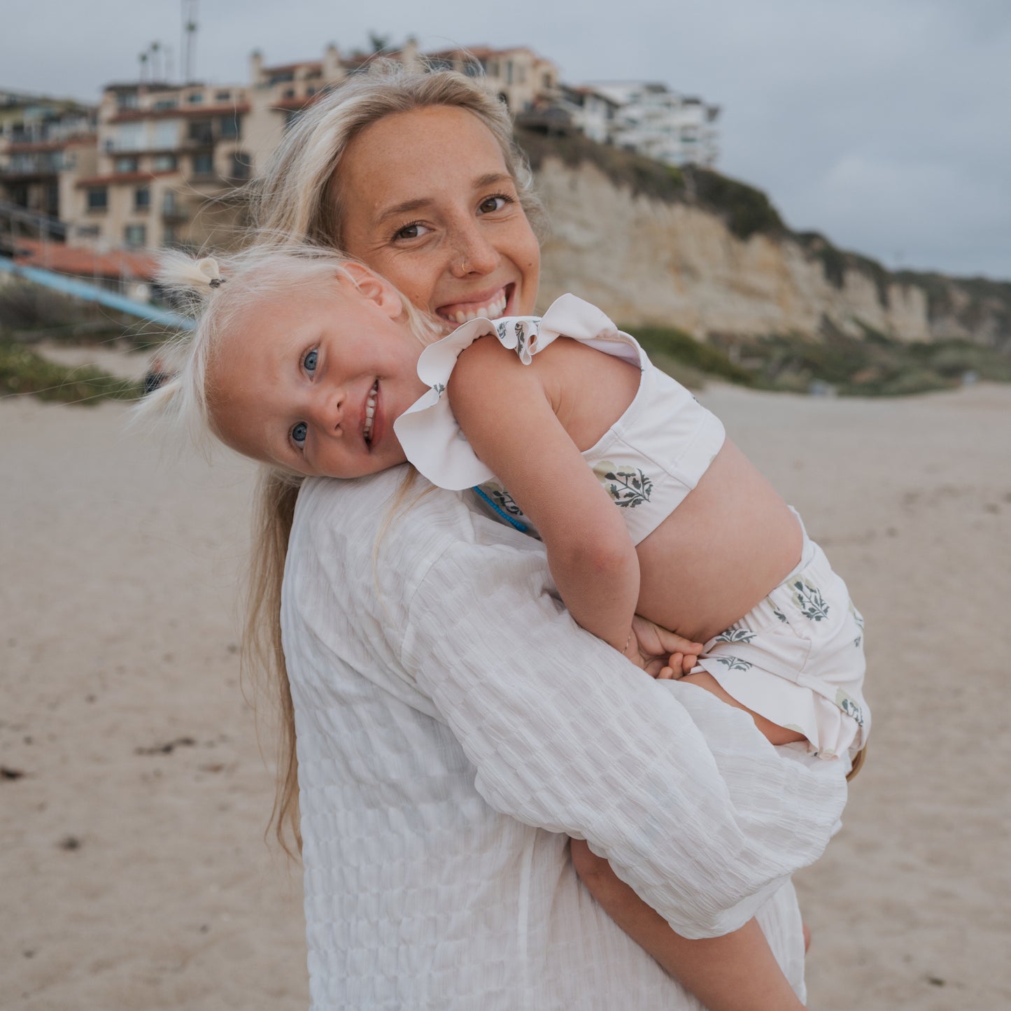 A smiling woman in a white dress holds a young girl with light blonde hair, wearing the forever french baby Girl's Two Piece Swimsuit | Marigold, on a sandy beach with cliffs and buildings in the background. Both look happy.