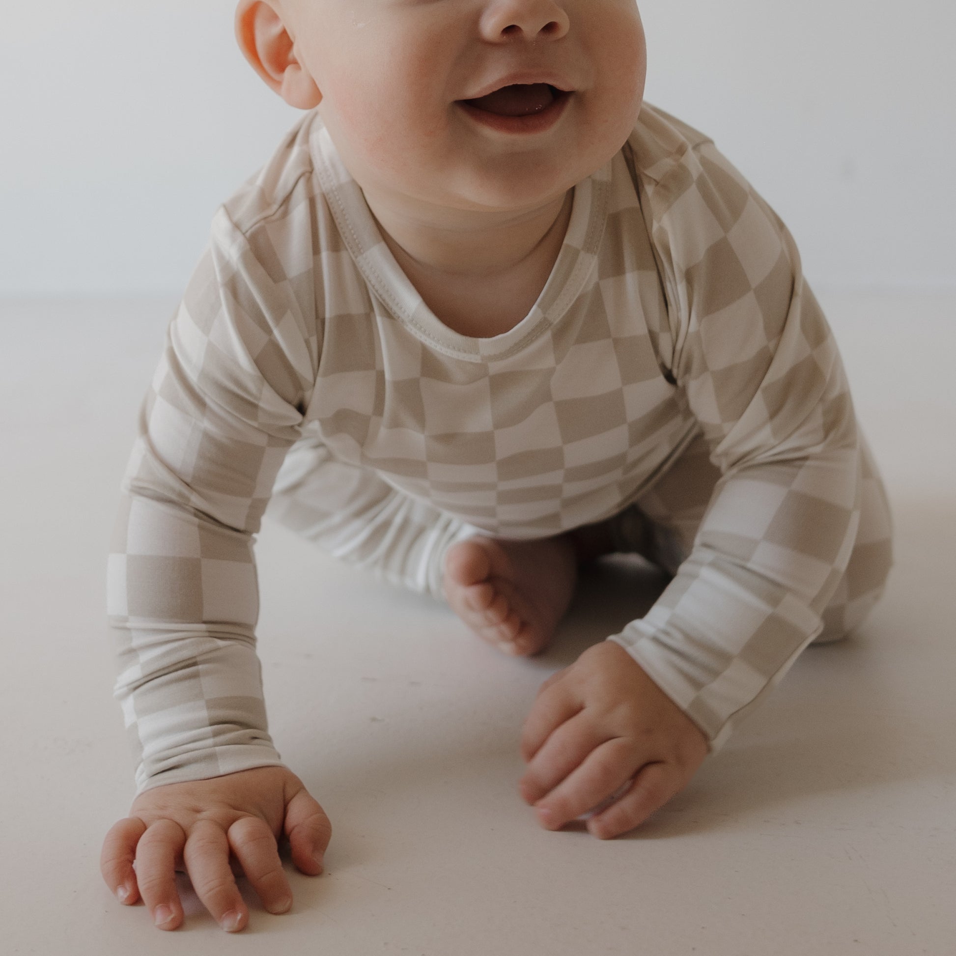 A smiling baby, lower face visible, crawls on a light floor wearing the forever french baby Child Sleepy Time Set | Checkerboard in beige and white.
