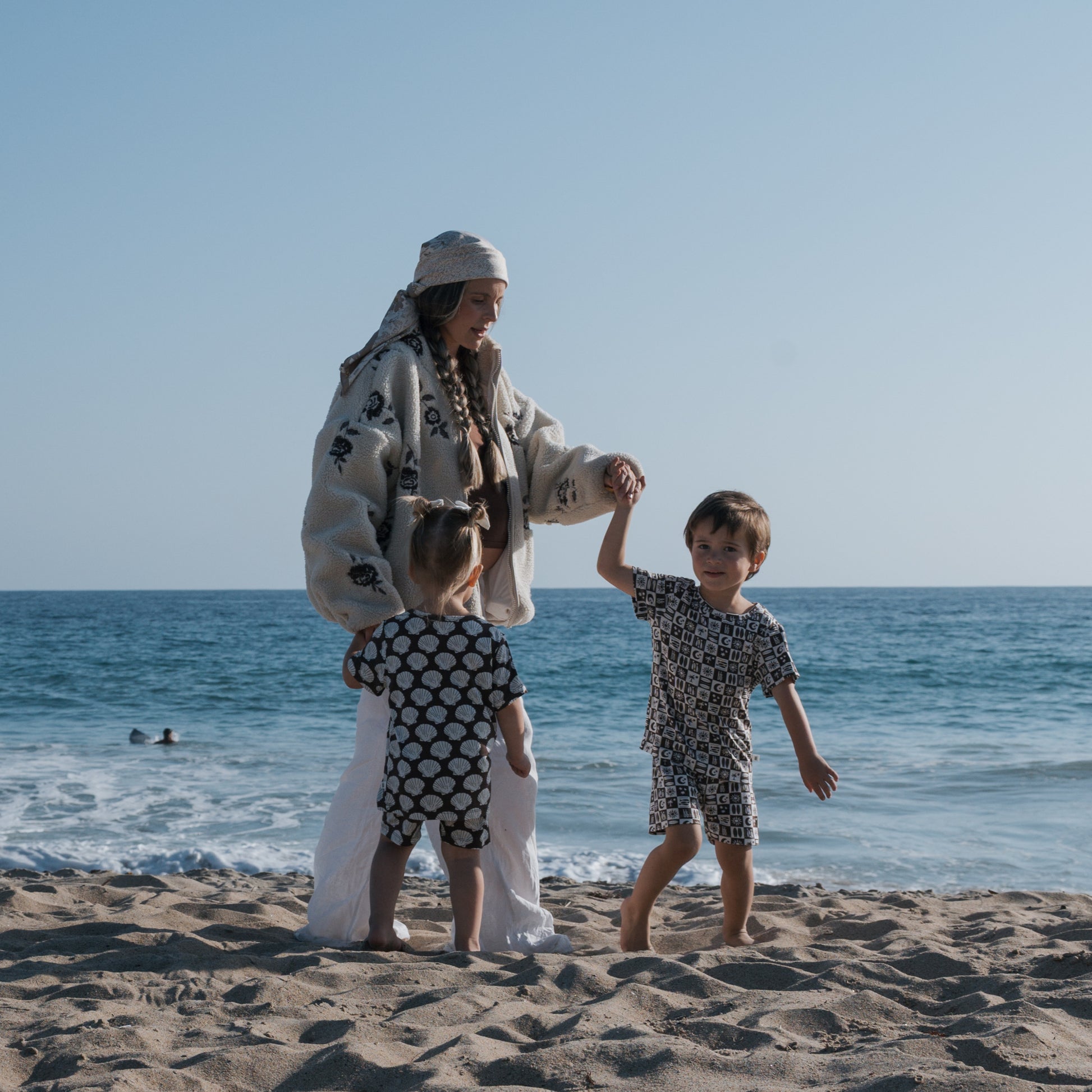 A woman in a headscarf and patterned jacket strolls a sandy beach, holding hands with two children dressed in forever french baby's Bamboo Short Set | Summer Check, with the ocean and clear sky behind them.