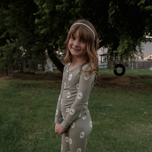 A young girl with long brown hair stands smiling on the grass in comfy forever french baby Bamboo Two Piece Pajamas | Mallard Frames, with trees and a tire swing in the background.
