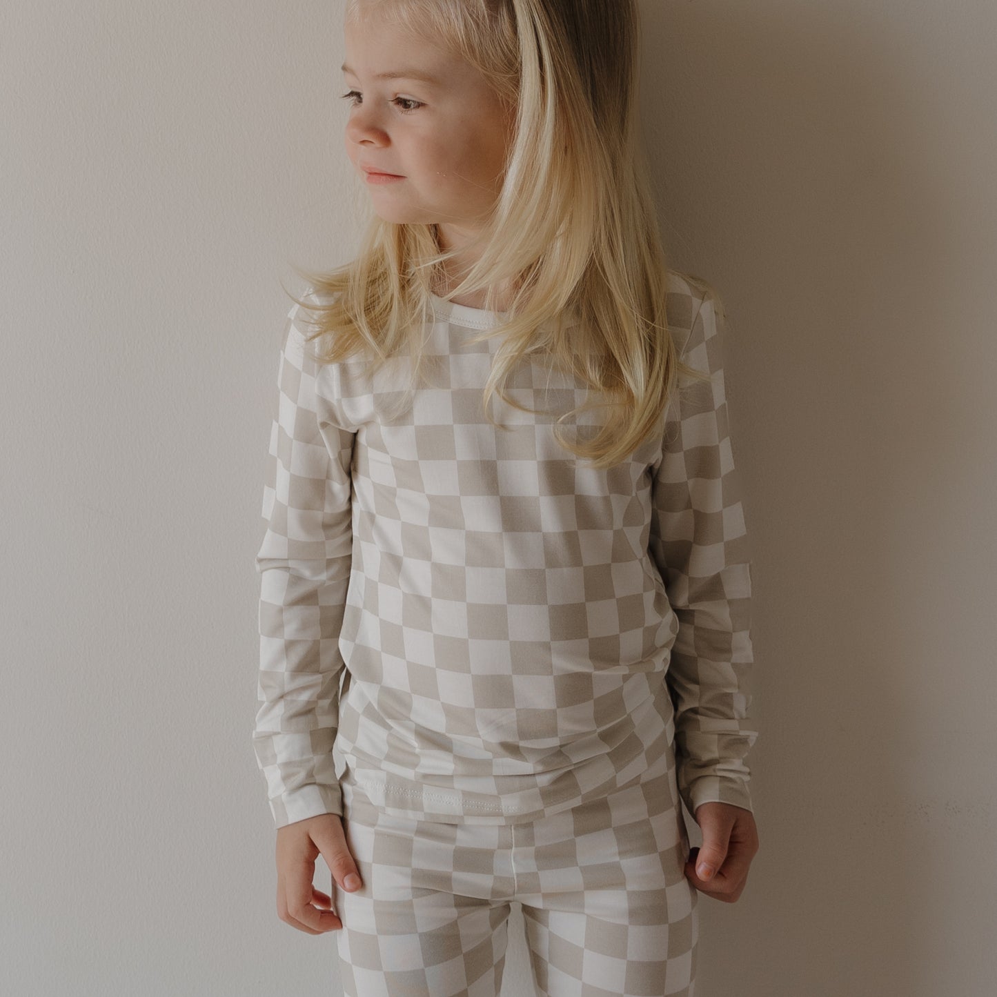 A young child with long blonde hair wears the forever french baby Child Sleepy Time Set | Checkerboard and stands against a plain light-colored wall, looking to the side and smiling softly.