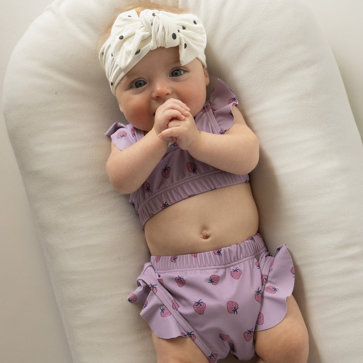 A baby wearing the forever french baby "Girl's Two Piece Swimsuit | Matilda" with a strawberry pattern and a dotted white headband lies on a soft white cushion, hands clasped in front of their mouth, gazing upward.