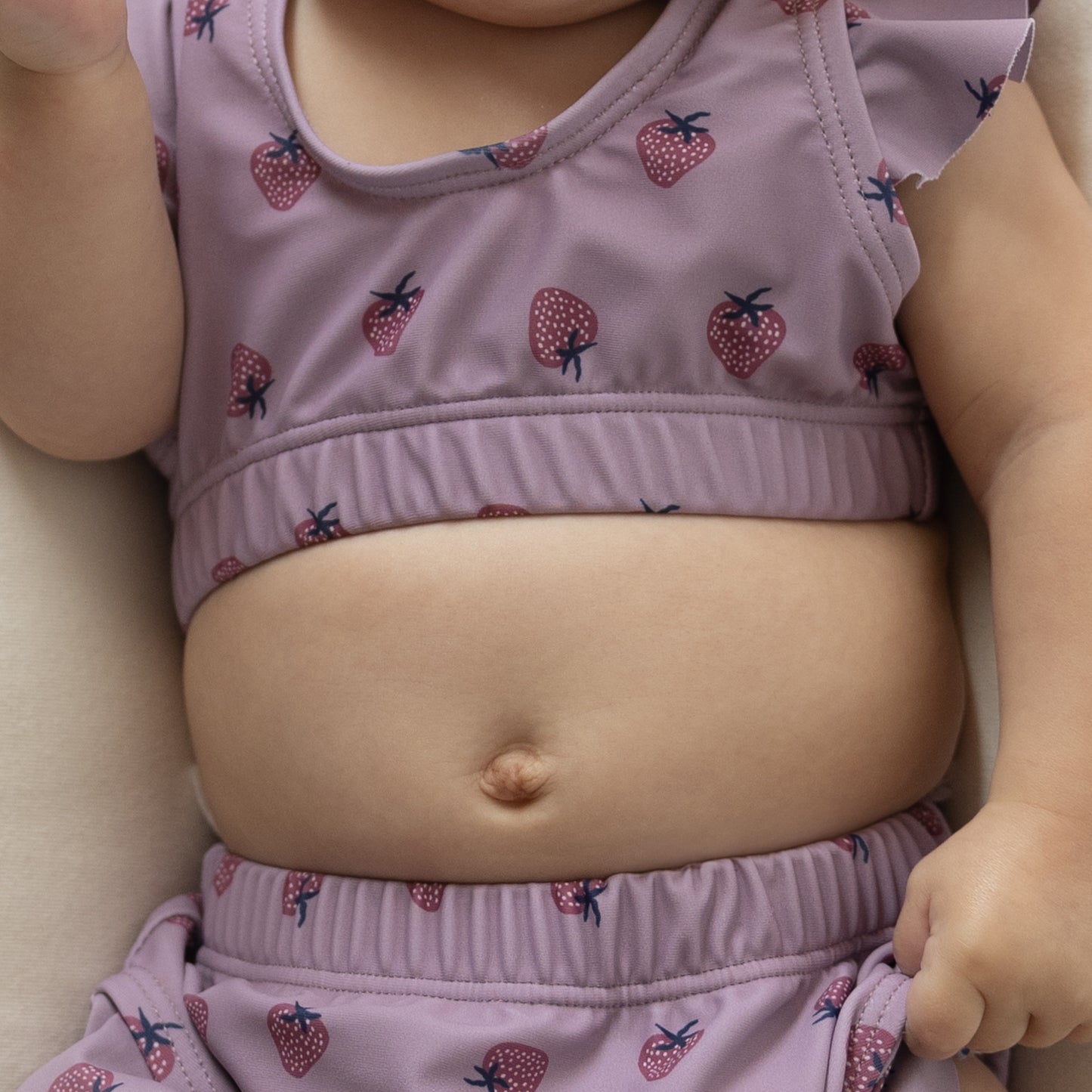 Close-up of a baby’s belly wearing the forever french baby Girl's Two Piece Swimsuit | Matilda, featuring a mauve shade with strawberry print. Only the torso, arm, and part of the leg are shown, highlighting skin and kids' swimwear details.
