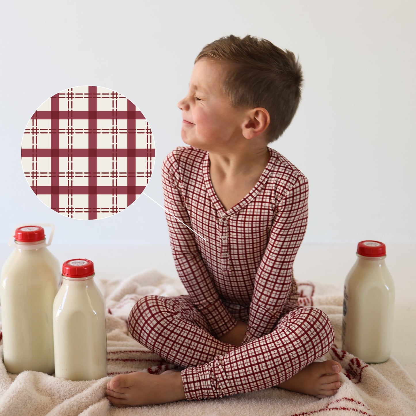 A young boy in forever french baby Bamboo Two Piece Pajamas | Window Pane sits cross-legged on a blanket by three glass milk bottles. A circular close-up highlights the pajama’s breathable fabric pattern beside him.