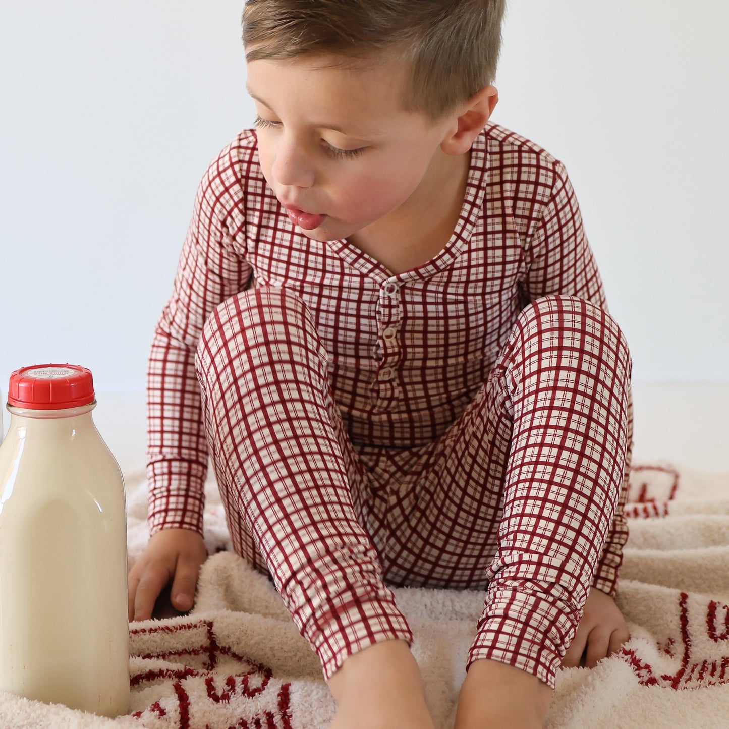 A young child wearing forever french baby's Bamboo Two Piece Pajamas | Window Pane sits on a blanket, gazing at a bottle of milk placed nearby.