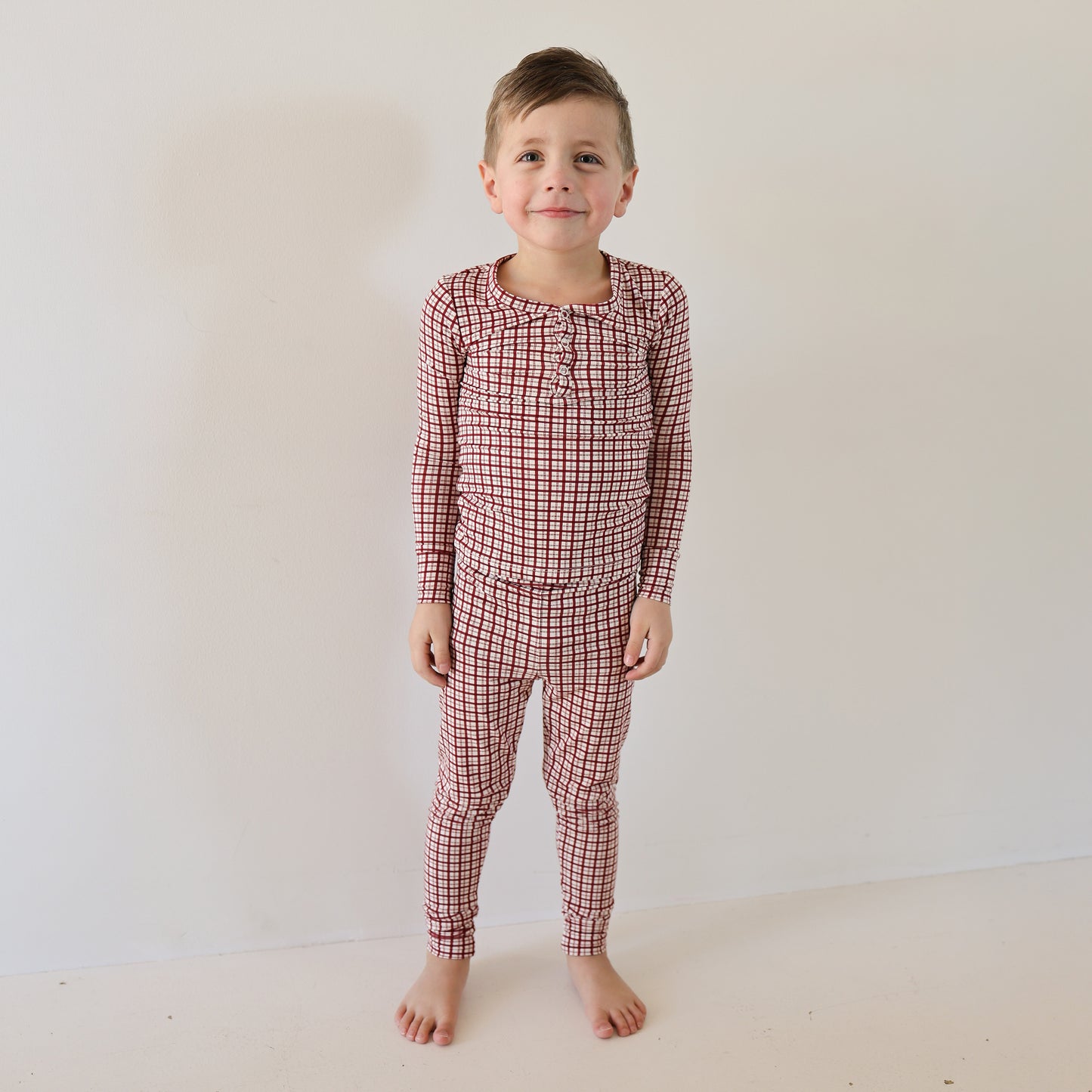 A young child stands barefoot against a white wall, wearing forever french baby’s Bamboo Two Piece Pajamas in Window Pane—soft, breathable sleepwear that brings comfort and an easy smile to bedtime.
