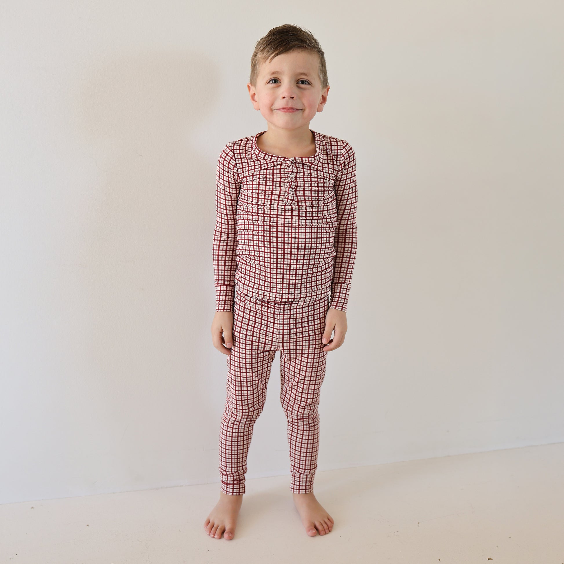 A young child stands barefoot against a white wall, wearing forever french baby’s Bamboo Two Piece Pajamas in Window Pane—soft, breathable sleepwear that brings comfort and an easy smile to bedtime.
