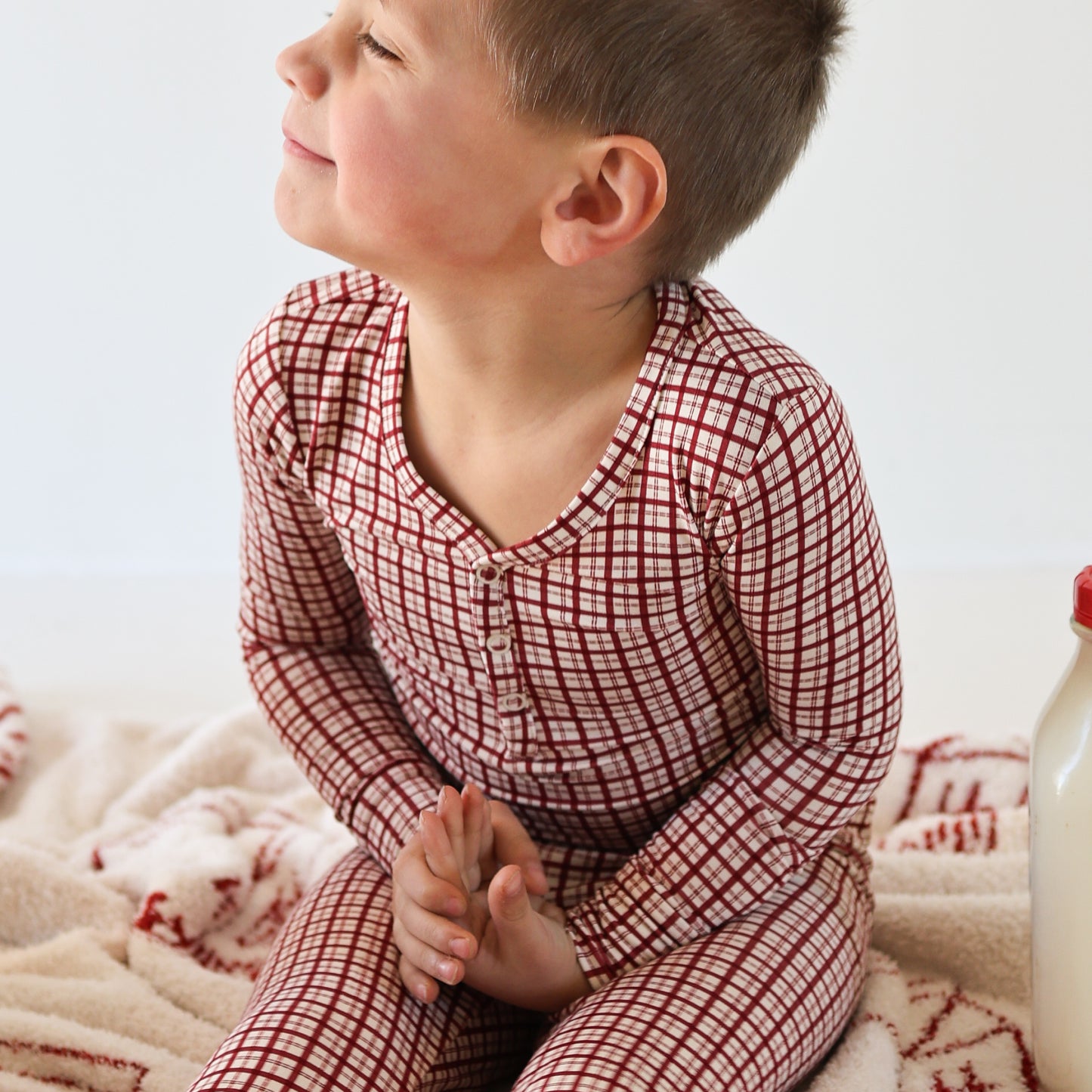 A young child in forever french baby Bamboo Two Piece Pajamas | Window Pane, sits cross-legged on a patterned blanket, smiling off to the side. A bottle is nearby, and the breathable sleepwear adds comfort to the minimal background.