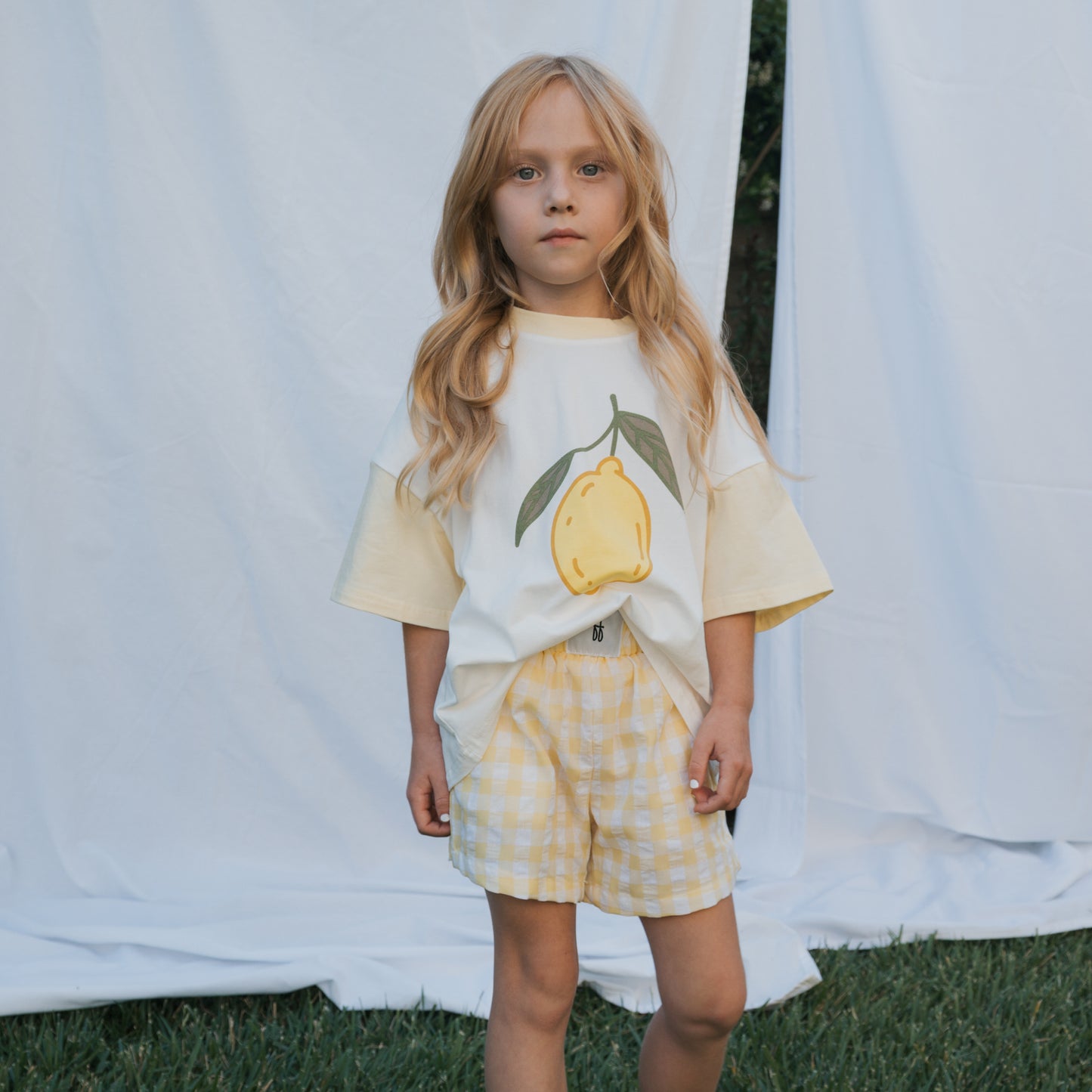 A young girl with long blonde hair stands on grass before a white backdrop, wearing the forever french baby Child Cotton Tee | Lemon and yellow gingham shorts—a charming summer look, gazing directly at the camera.