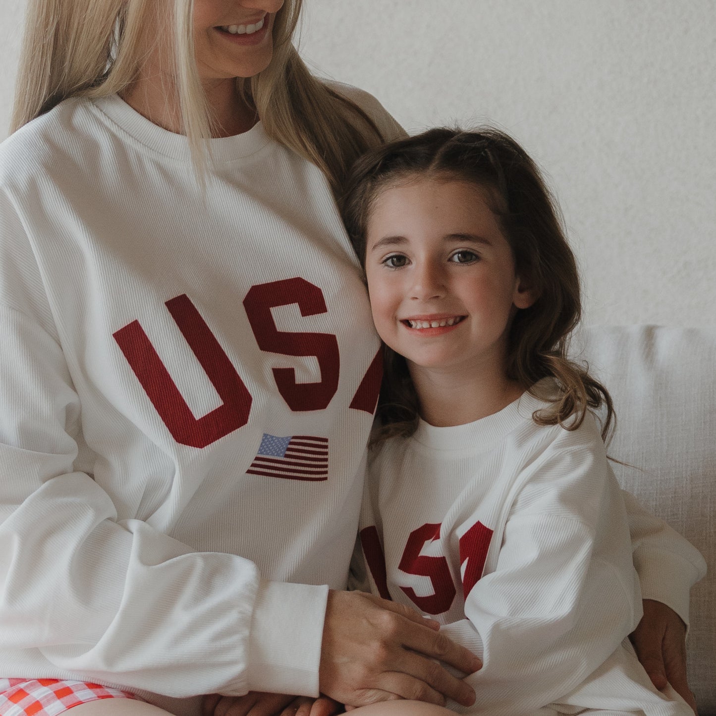 A woman and a girl sit together in matching "USA" *PRE-ORDER* Adult Sweatshirts by forever french baby, both featuring an American flag. The girl's smile and their relaxed fit add comfort to the sweet moment.