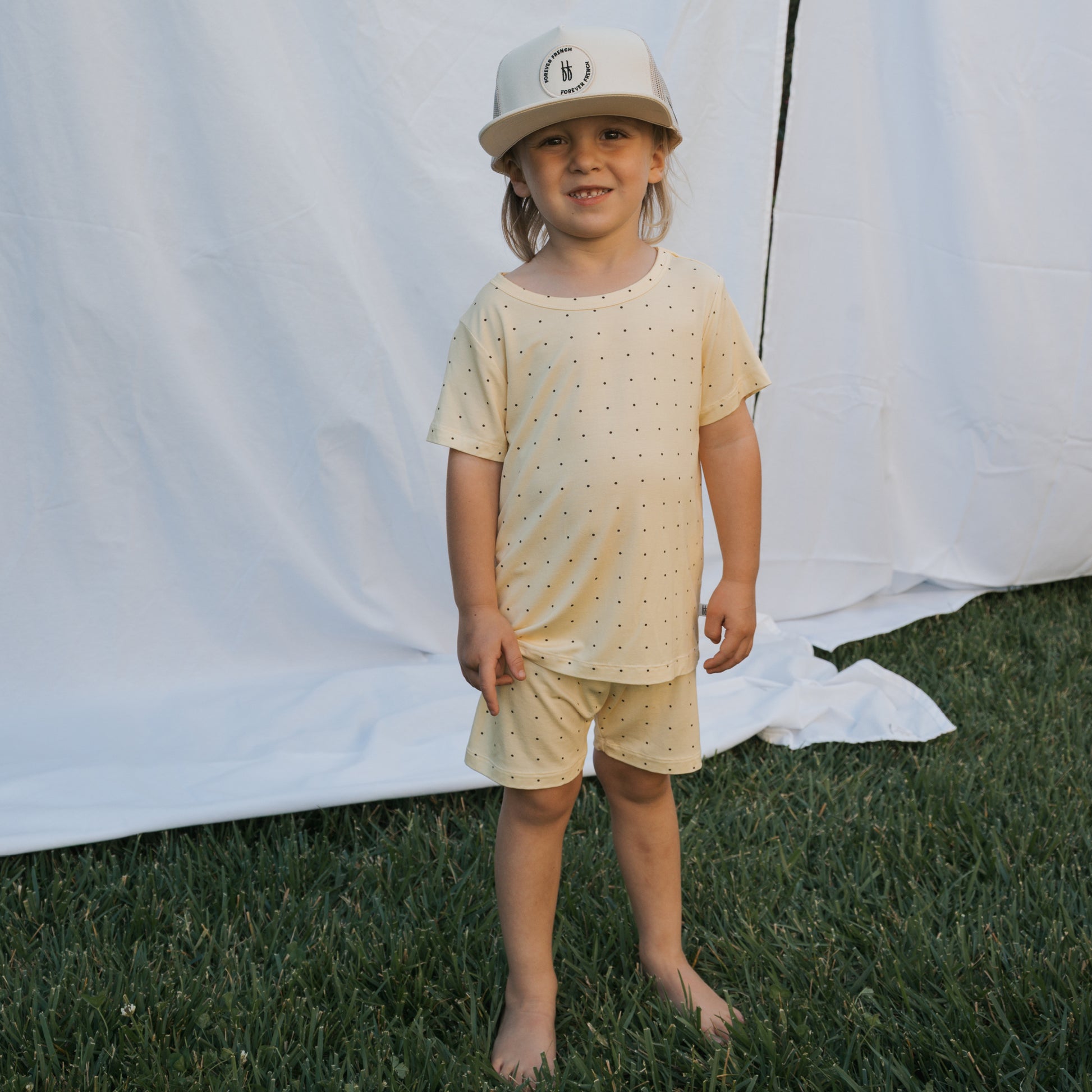 A young child stands barefoot on grass in the forever french baby Bamboo Short Set | Veda—breathable, light-colored polka dot shorts and t-shirt with a light baseball cap—posed outdoors before a white sheet backdrop.