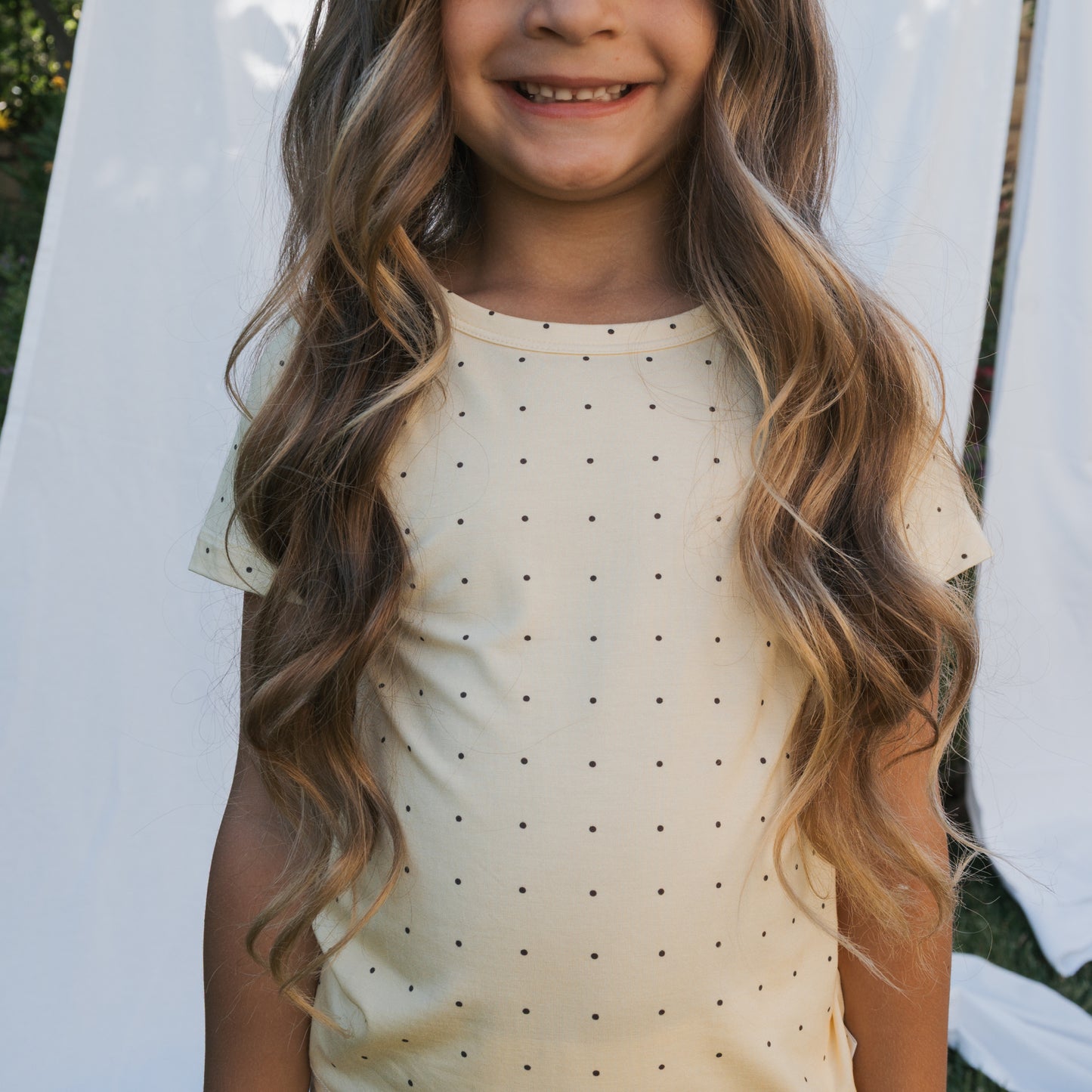 A young girl in the forever french baby Bamboo Short Set | Veda—a cream short-sleeved top with black polka dots—smiles outdoors in front of white fabric, showcasing stylish and breathable children’s clothing.