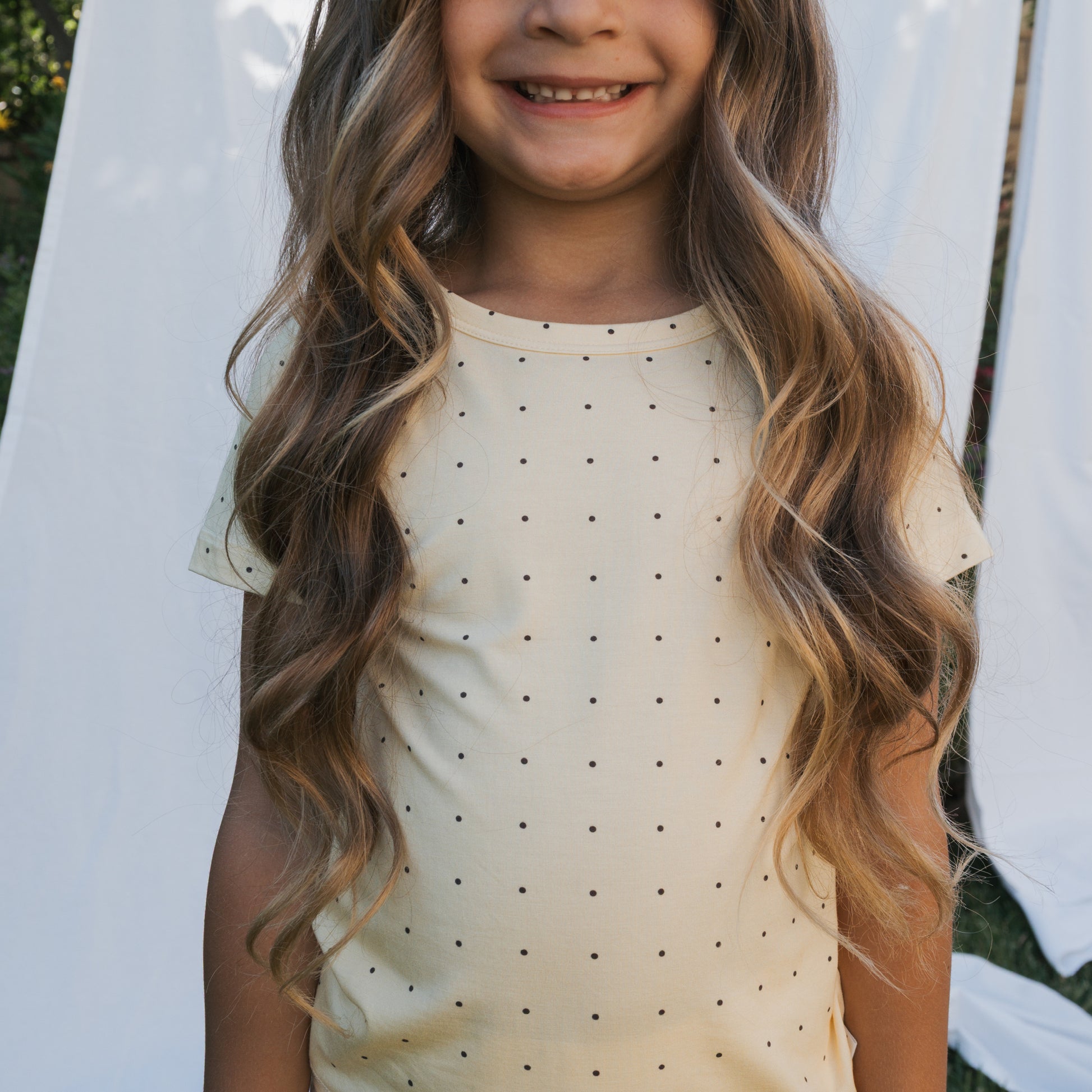 A young girl in the forever french baby Bamboo Short Set | Veda—a cream short-sleeved top with black polka dots—smiles outdoors in front of white fabric, showcasing stylish and breathable children’s clothing.