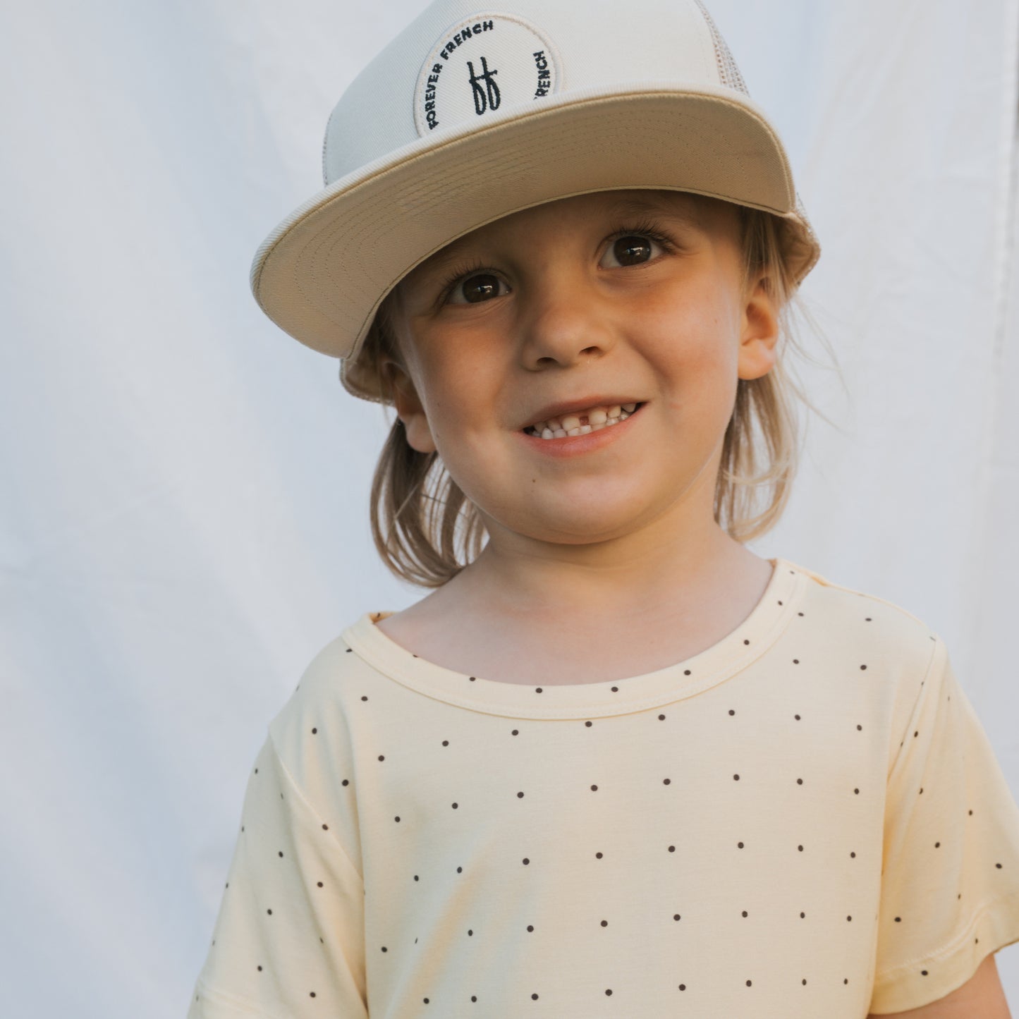 A young child with long hair smiles warmly in front of a light background, wearing the breathable Bamboo Short Set | Veda by forever french baby—a cream-colored, polka-dotted outfit and matching hat.
