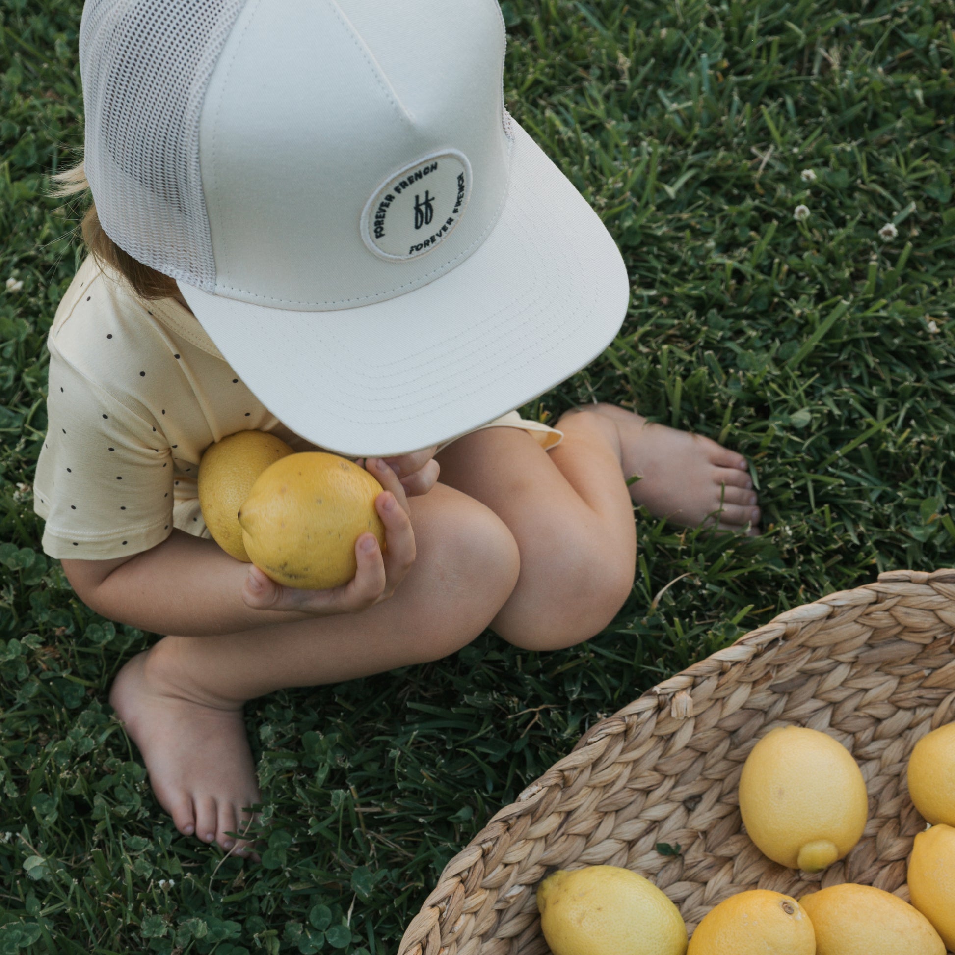 A young child in the forever french baby Bamboo Short Set | Veda—wearing a white cap and yellow, dotted shirt—sits barefoot on grass holding two lemons beside a woven basket filled with lemons.