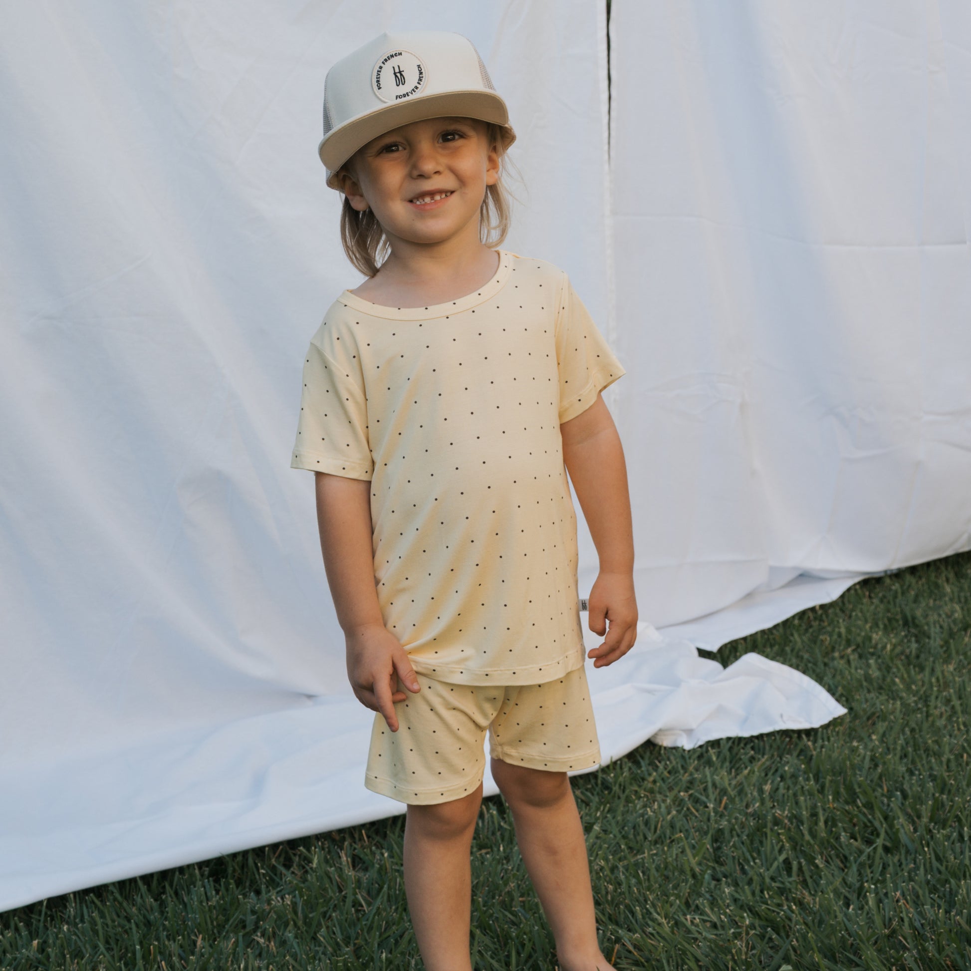 A young child smiles on grass before a white fabric backdrop, wearing the forever french baby Bamboo Short Set | Veda—a breathable cream shirt and shorts with black dots—and a beige logo cap.