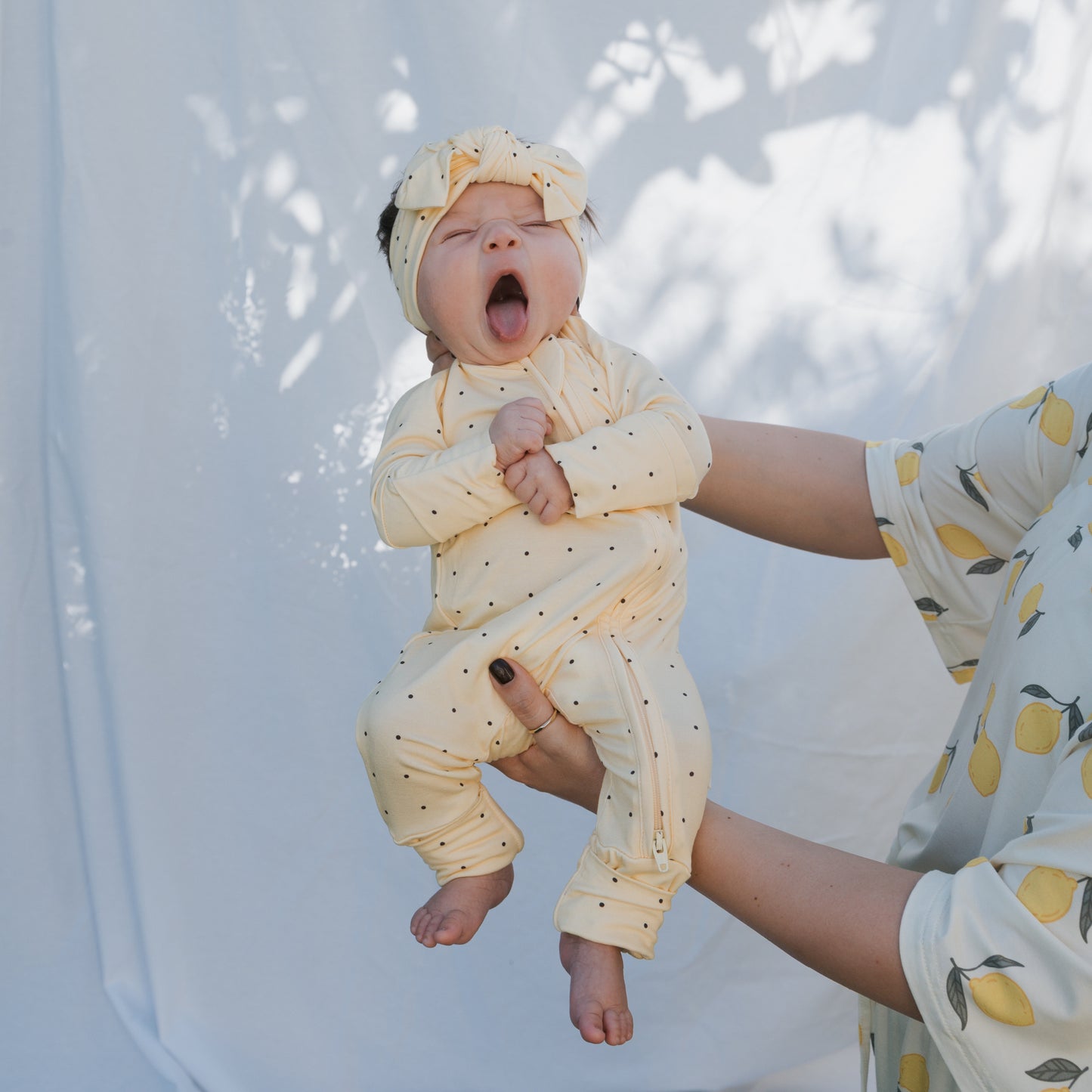 A baby in forever french baby Bamboo Zip Pajamas | Veda, featuring yellow polka dots and a matching headband, yawns while held by an adult in a lemon-print shirt against a sunlit white background.