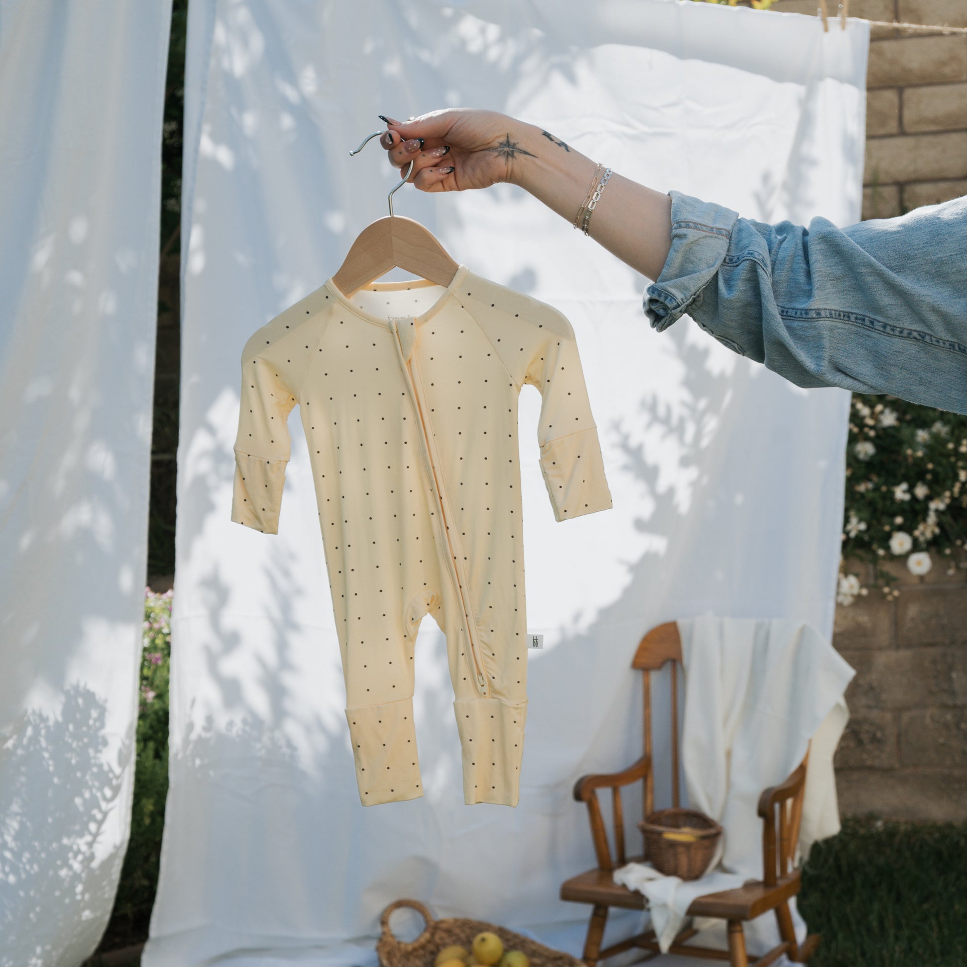 Someone holds up the forever french baby Bamboo Zip Pajamas | Veda, a yellow, polka-dotted breathable onesie, on a hanger outdoors. Behind them are a wooden rocking chair with a white blanket and a basket of apples by white sheets and stone wall.