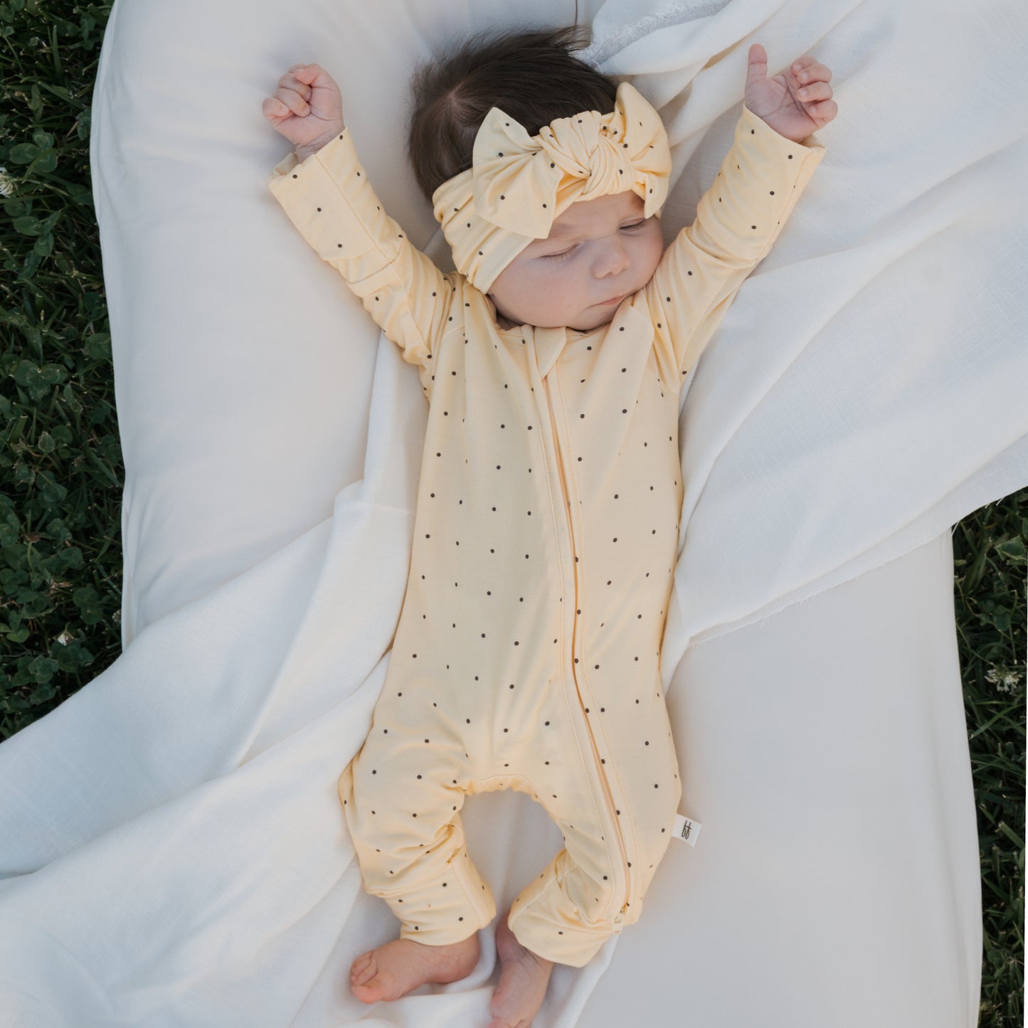 A baby in forever french baby's Bamboo Zip Pajamas | Veda—a yellow, polka-dotted onesie with a matching headband—sleeps peacefully on a white blanket with arms outstretched, lying outdoors on green grass.