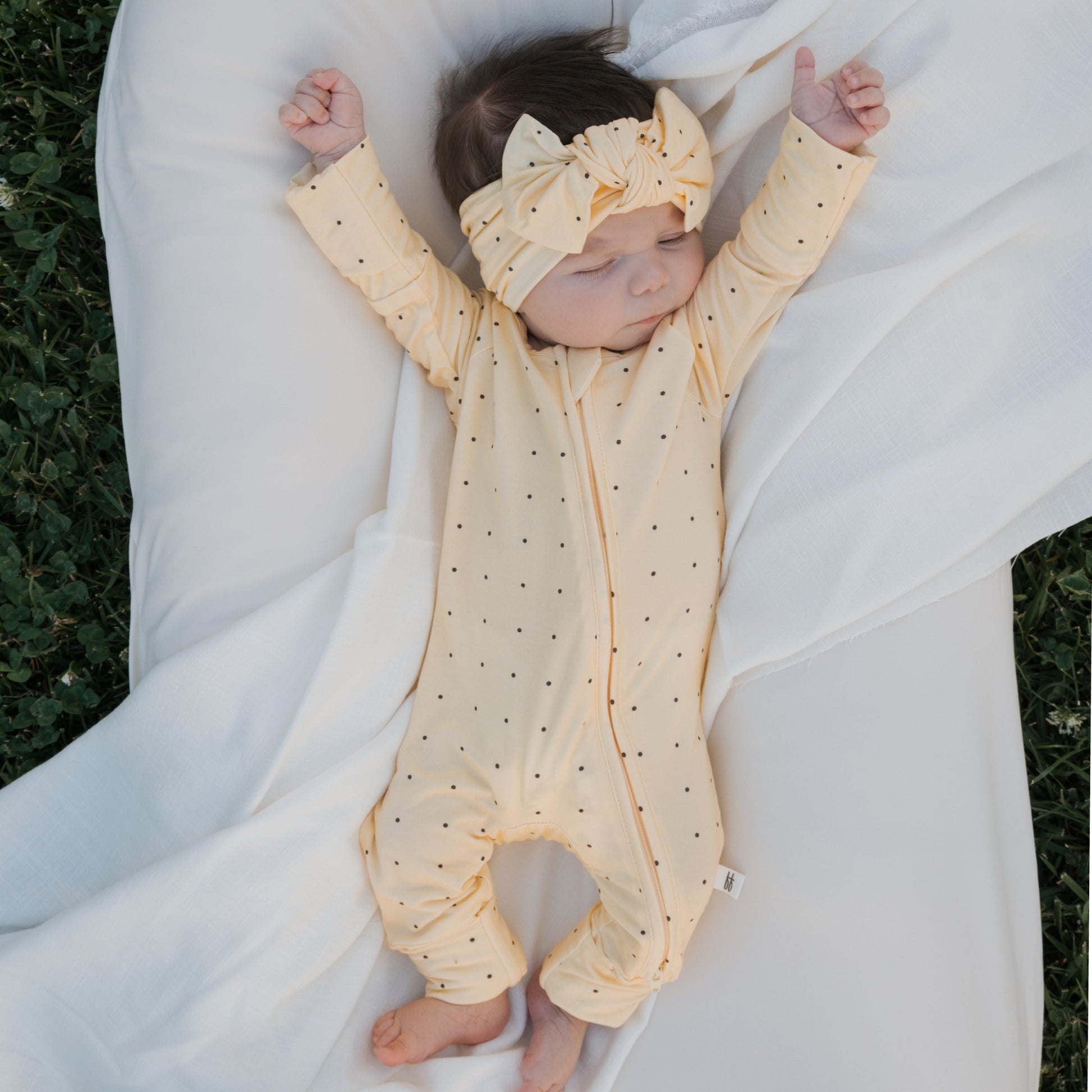 A baby in forever french baby's Bamboo Zip Pajamas | Veda—a yellow, polka-dotted onesie with a matching headband—sleeps peacefully on a white blanket with arms outstretched, lying outdoors on green grass.