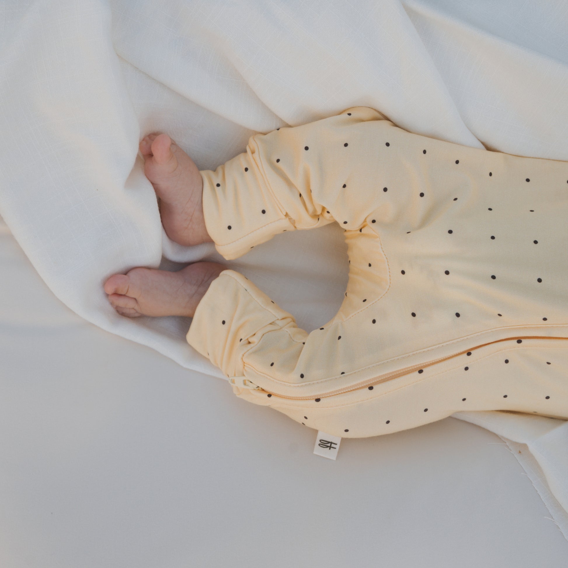 A baby lies on a white sheet, legs showing. They wear forever french baby's Bamboo Zip Pajamas | Veda in light yellow with black dots—breathable bamboo sleepwear for comfort. One foot is partly tucked under the pants.