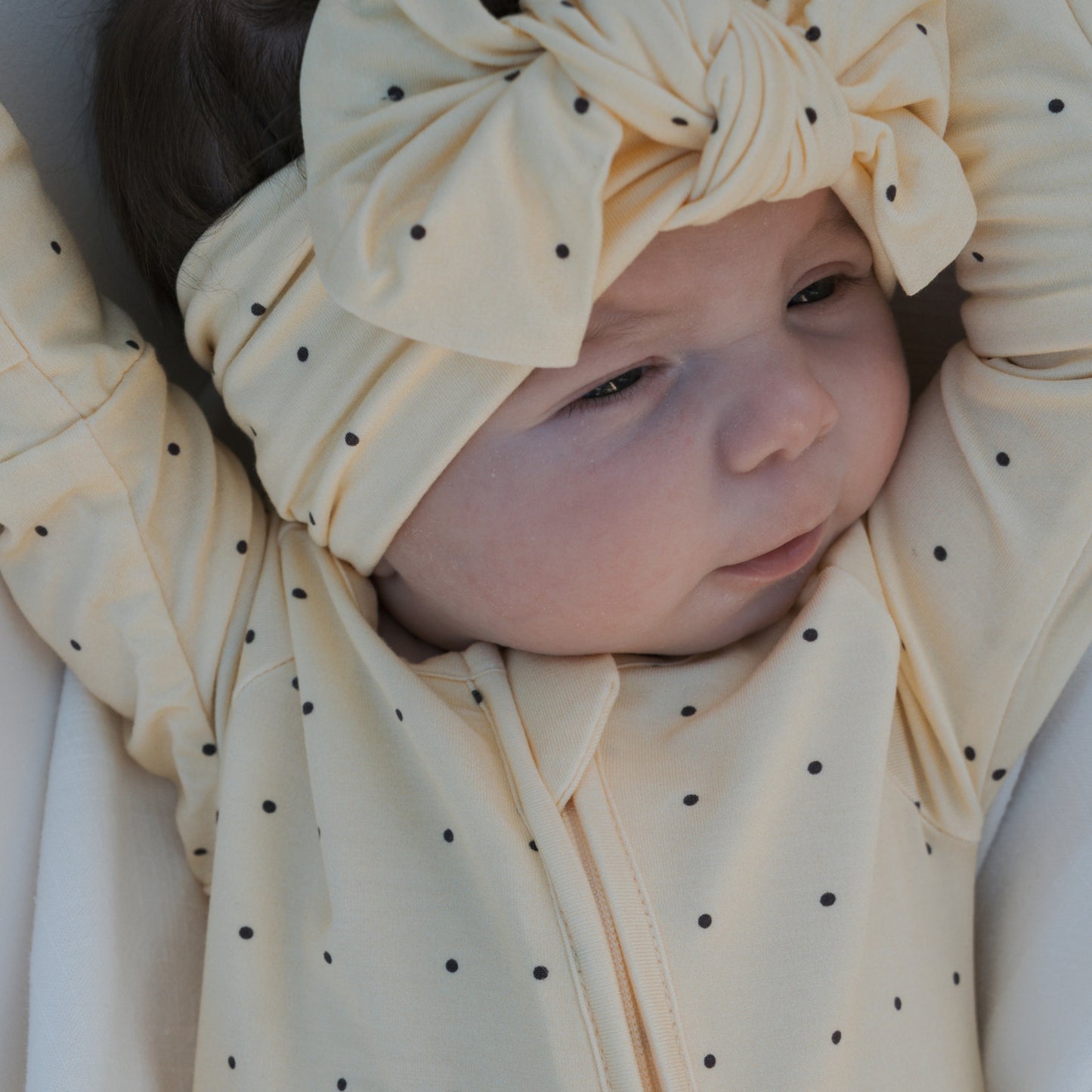 A baby in pale yellow forever french baby Bamboo Zip Pajamas | Veda with black polka dots and a matching large-bow headband lies down with arms raised.
