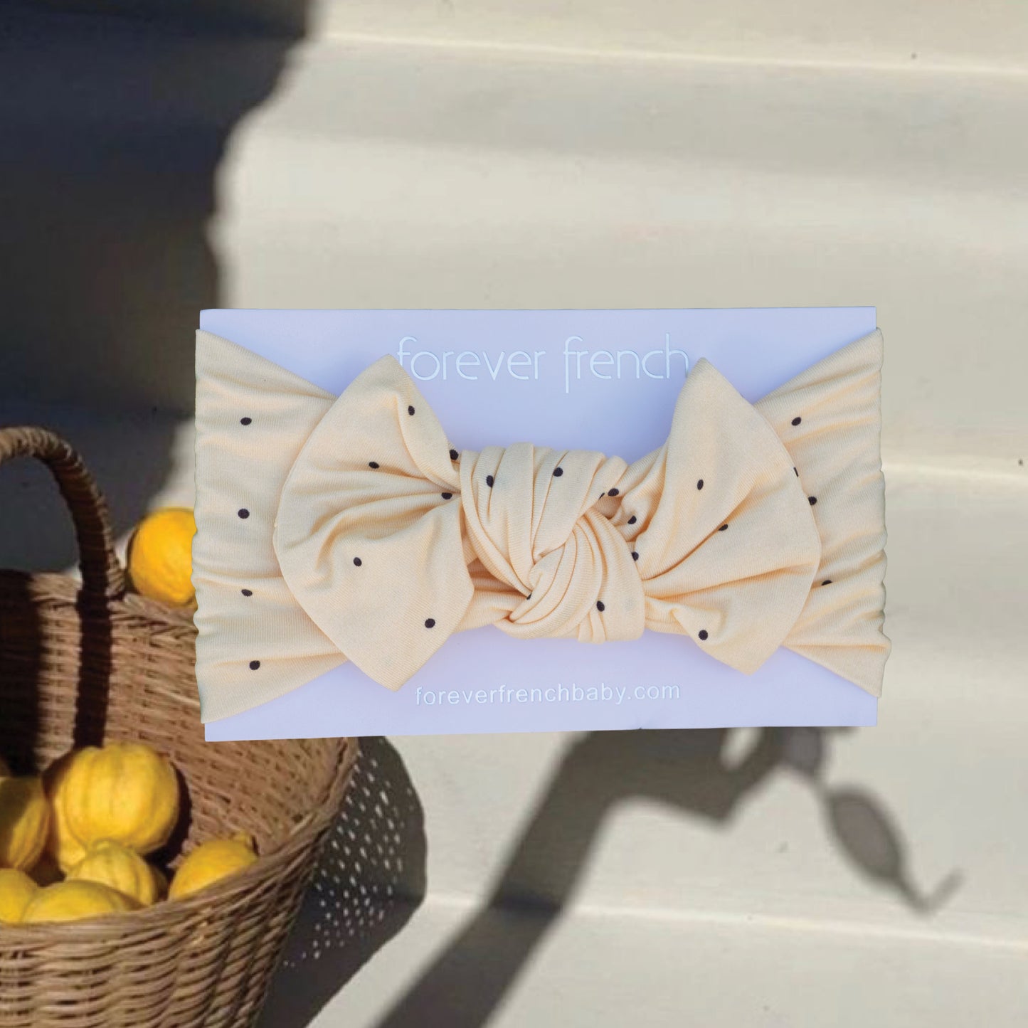 The Bamboo Head Wrap | Veda by forever french baby, cream-colored with black polka dots and a large bow, is shown on a light blue background beside a basket of small yellow fruits on sunlit steps.