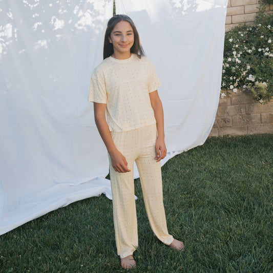 A young person stands barefoot on grass, wearing the forever french baby Child Boxy Tee Lounge Set | Veda—light-colored, breathable bamboo clothing with small dots. A white sheet, brick wall, and flowering bushes form the background.