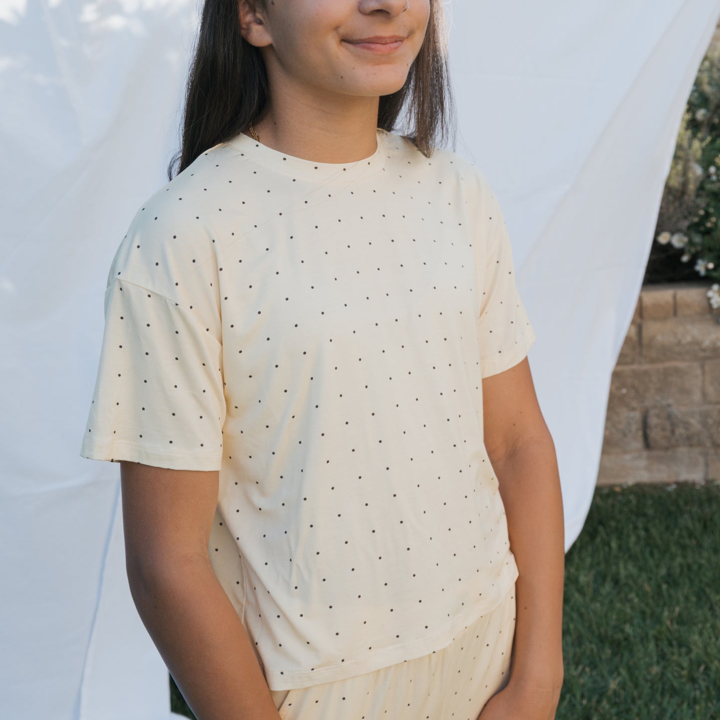 A person stands outdoors in front of a white sheet backdrop, wearing the forever french baby Child Boxy Tee Lounge Set | Veda. They have long dark hair, are smiling slightly with hands in front. Grass and a stone wall are visible.